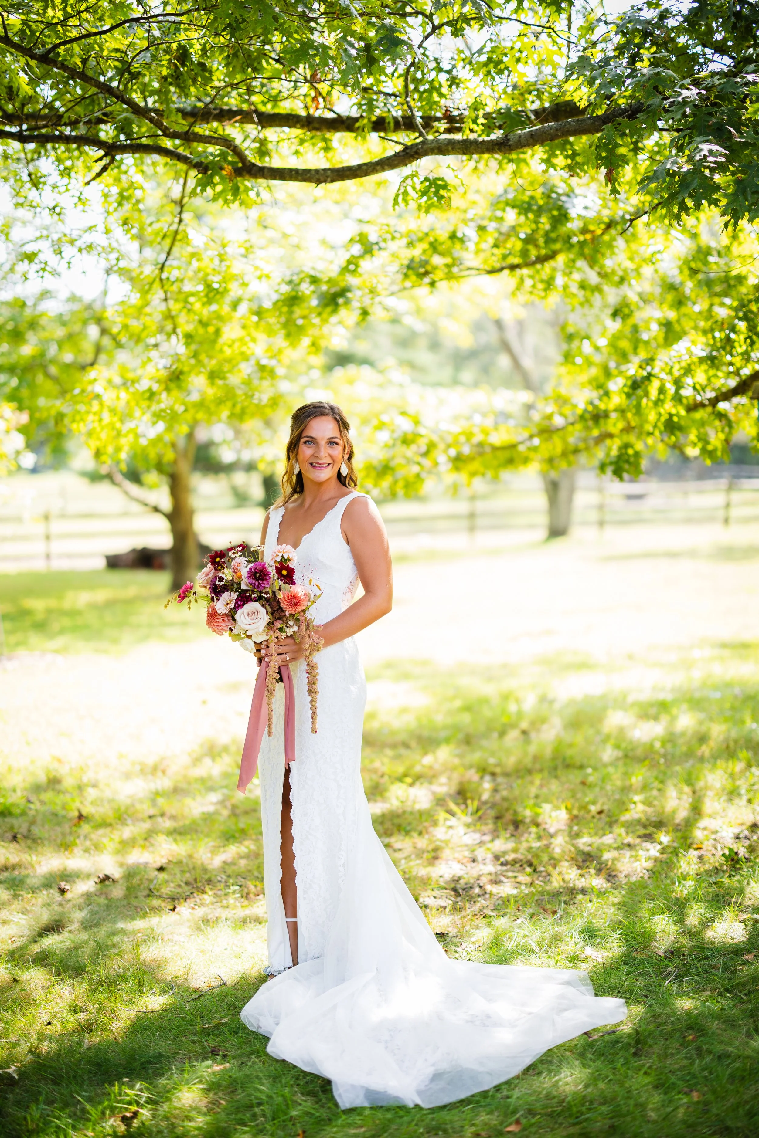 A woman in a white wedding dress holding a bouquet of flowers outdoors in a grassy area with trees in the background.