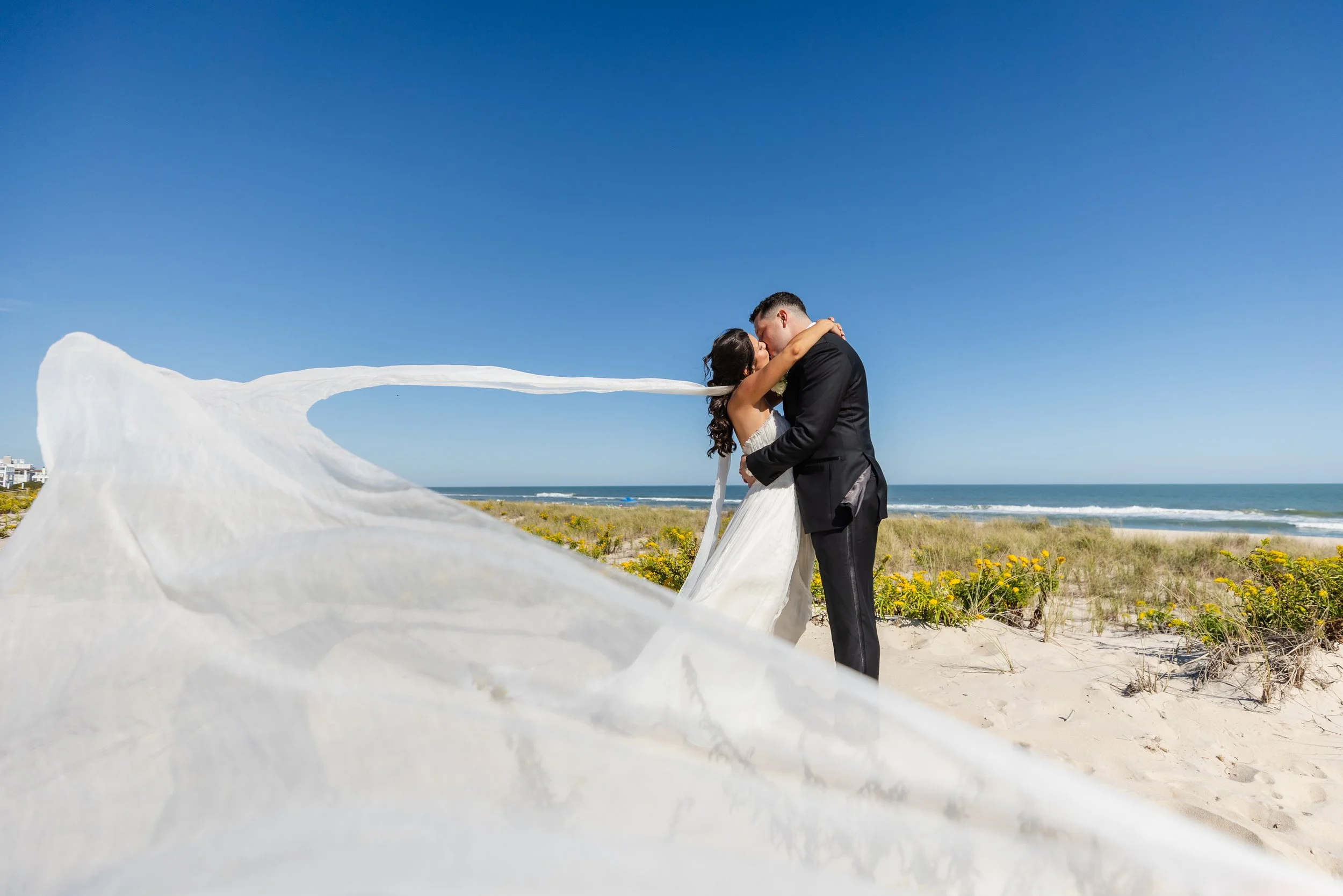 A bride and groom embrace and kiss on a beach, with the bride's veil flowing in the wind and the ocean in the background.