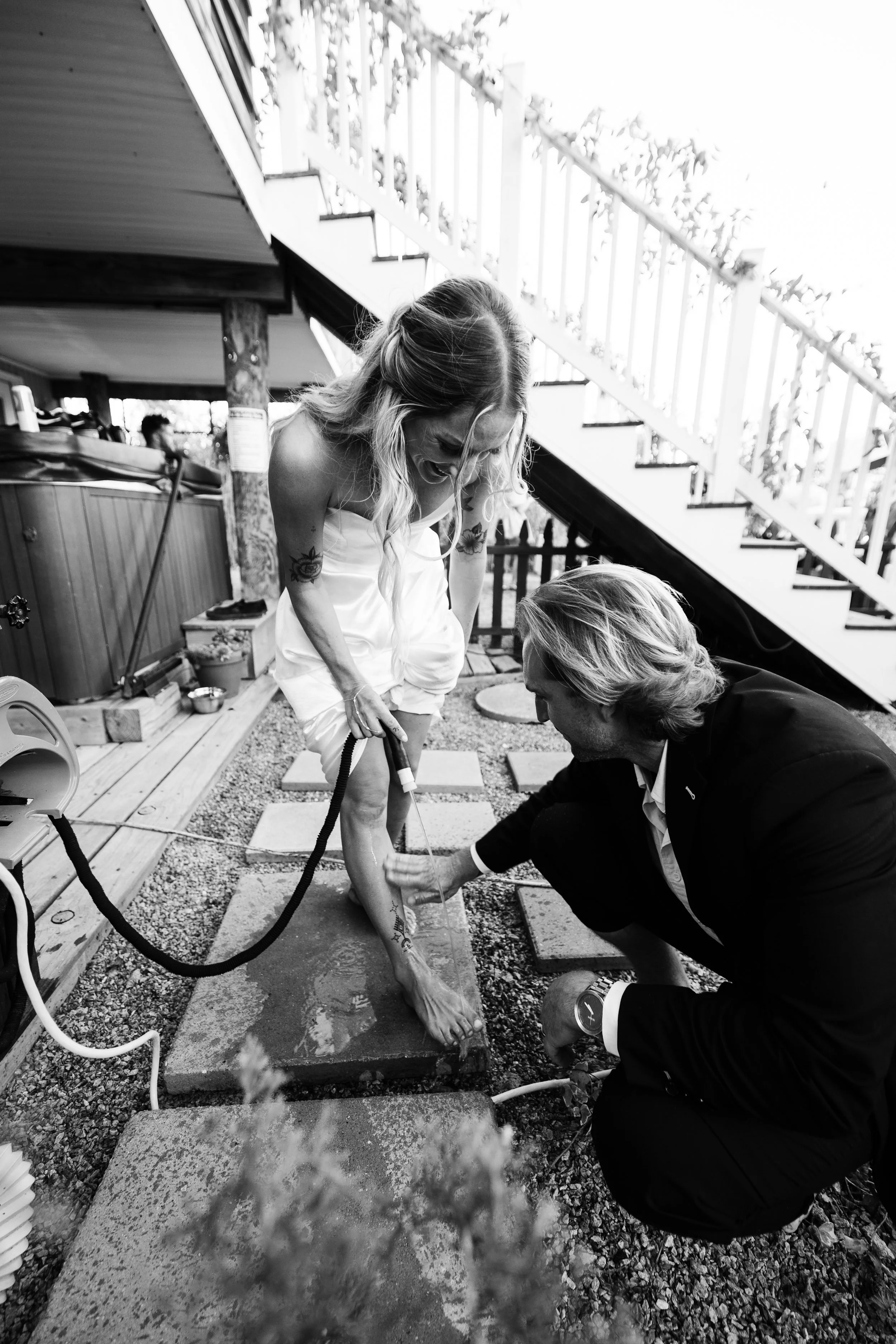 A woman in a white dress getting a tattoo on her lower leg while a man in a black suit supervises.
