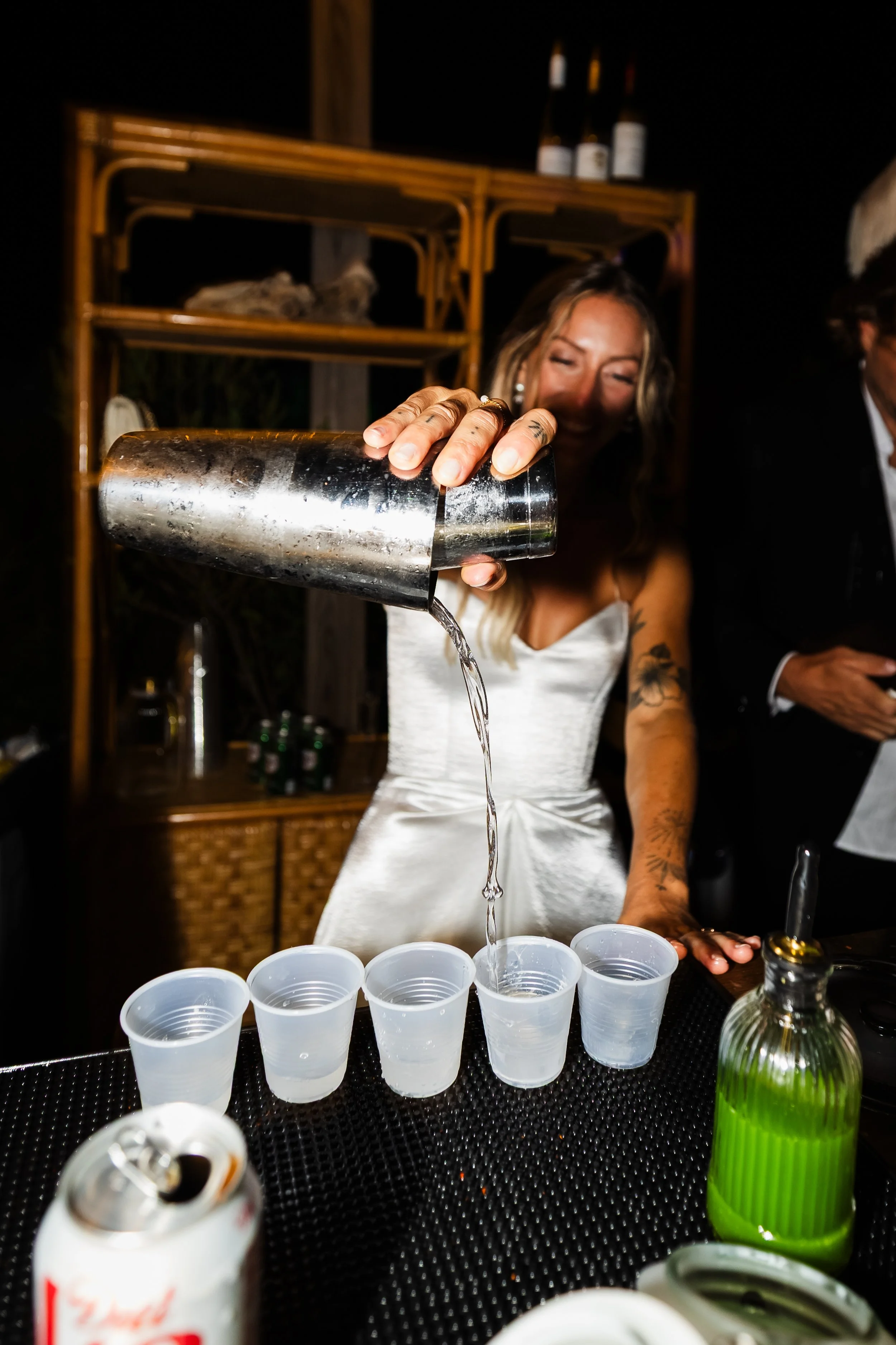 A woman in a white dress pouring liquid into small plastic cups at a bar or party setting.