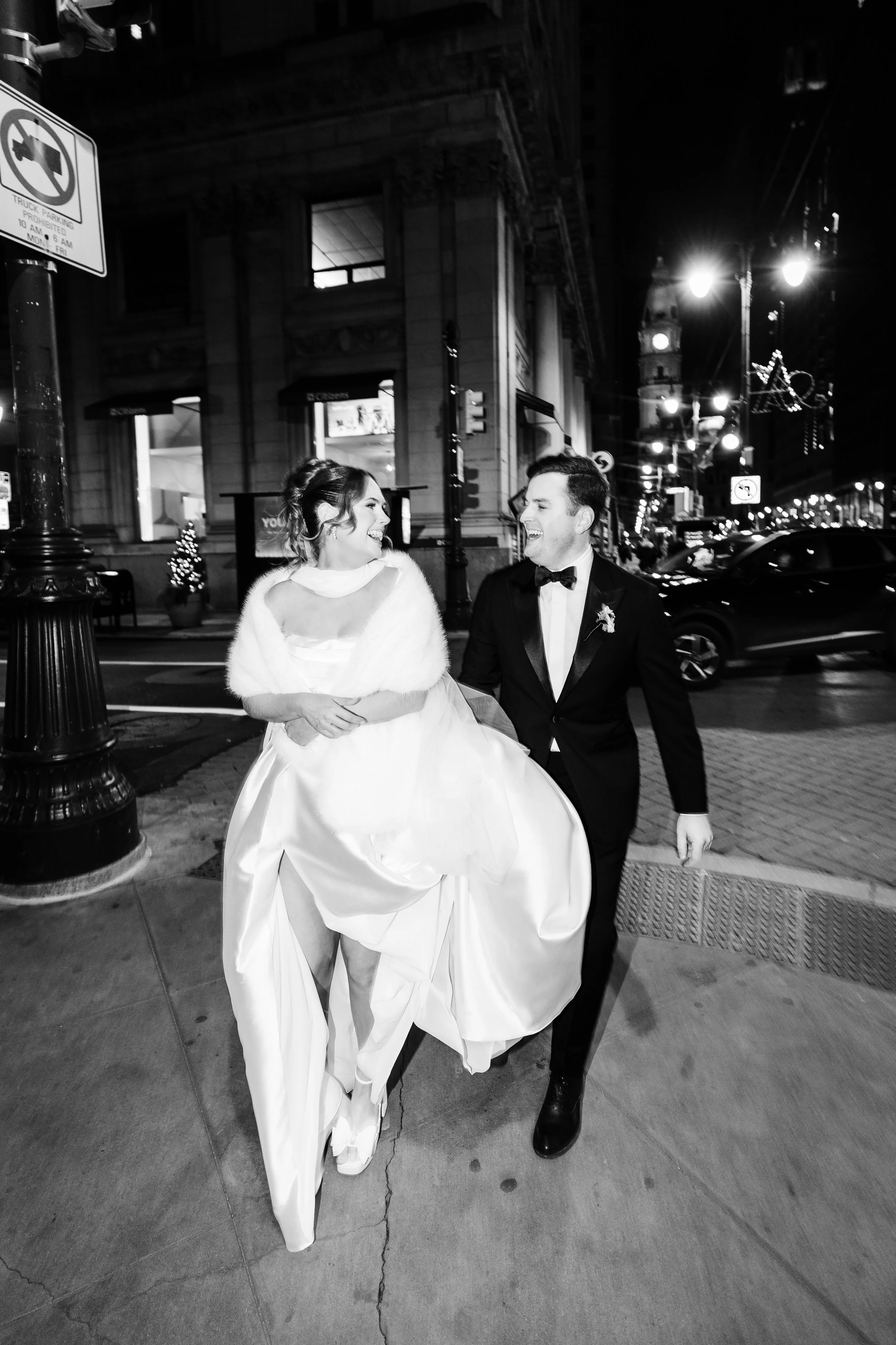 A bride and groom walking on a city sidewalk at night, smiling and looking at each other, with streetlights and buildings in the background.