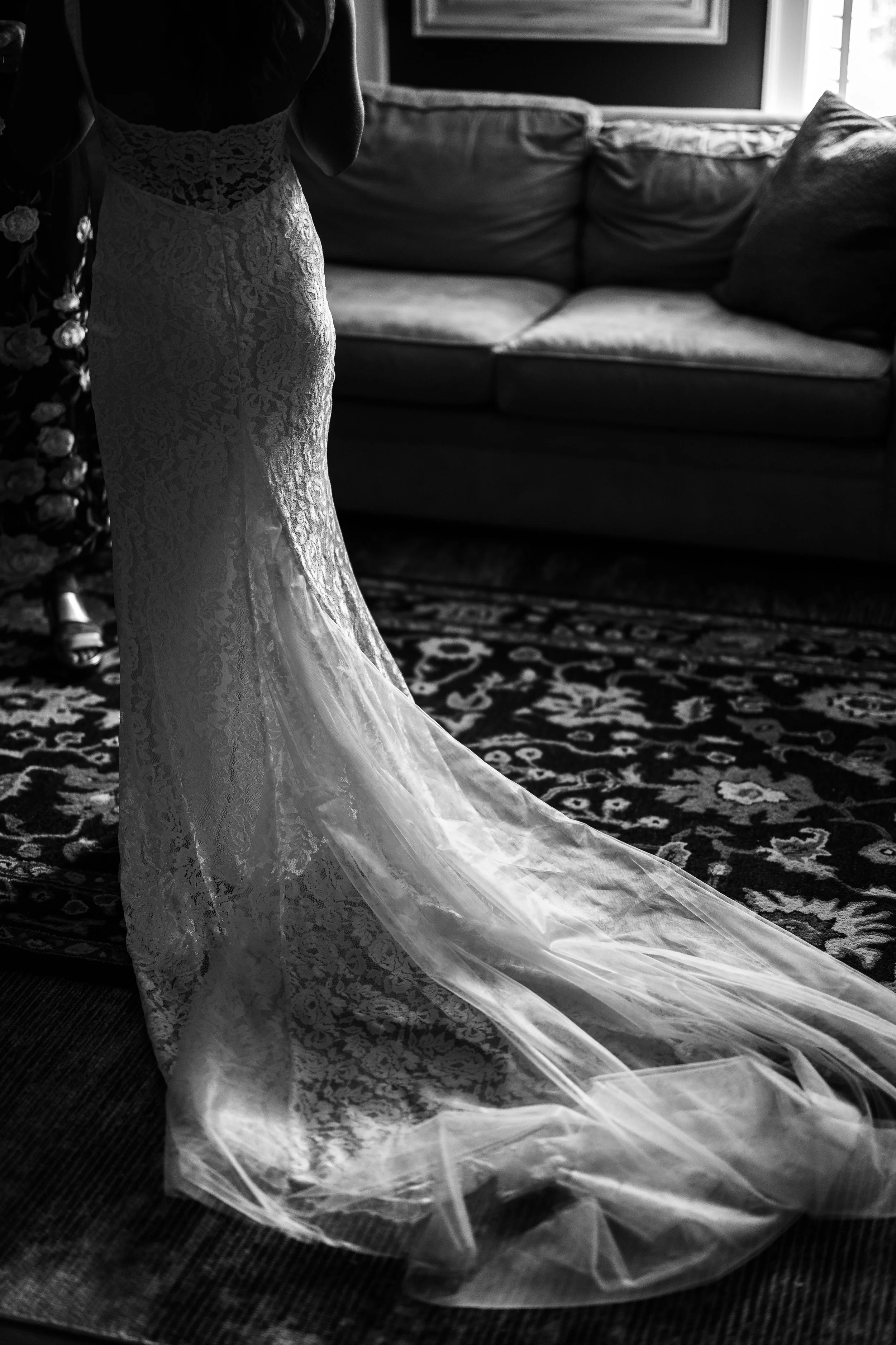 A woman in a lace wedding dress standing on a patterned rug indoors, with a sofa and window in the background.
