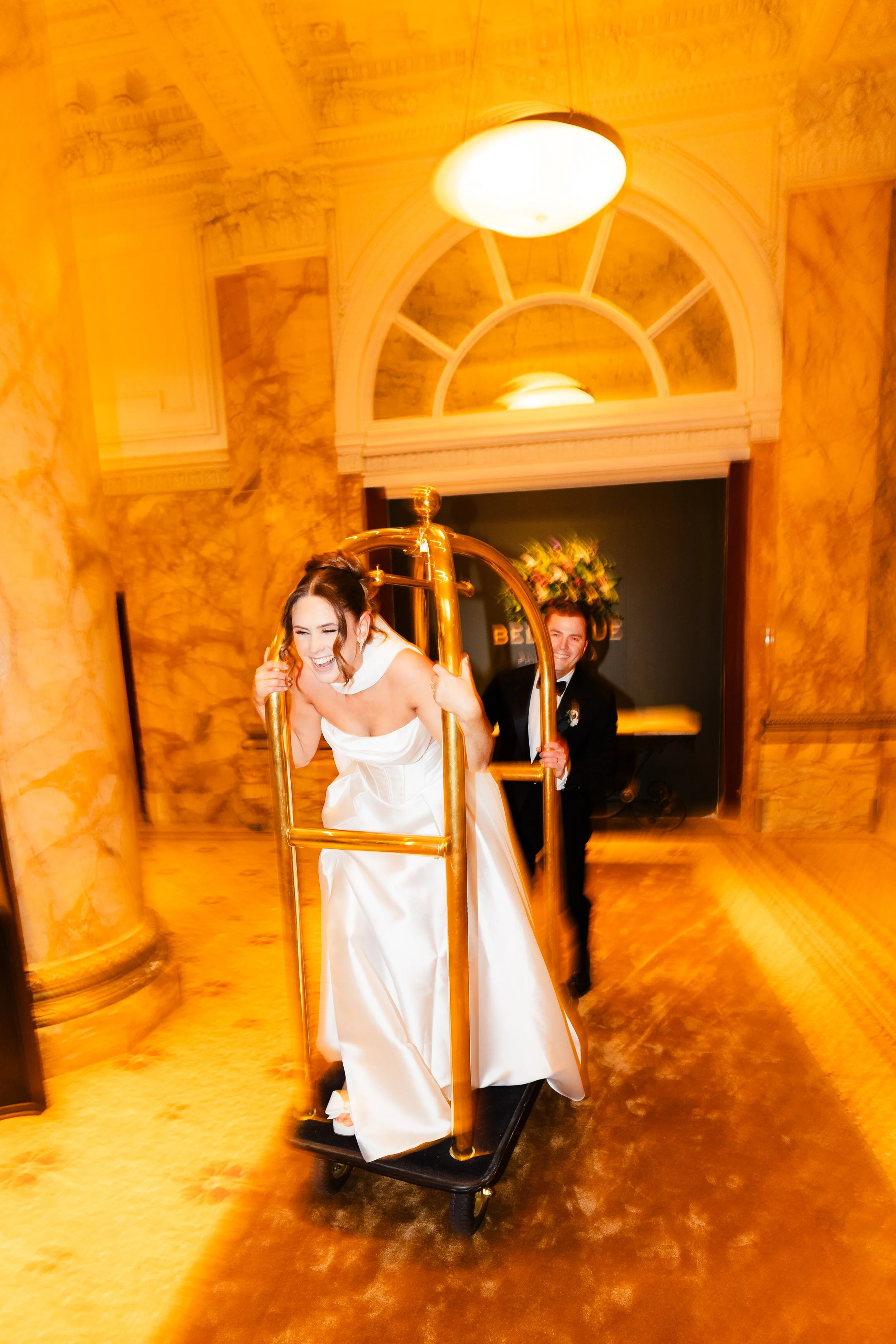 Bride in a white wedding dress laughing on a luggage cart with groom dressed in a tuxedo pushing behind her in an ornate hotel lobby.