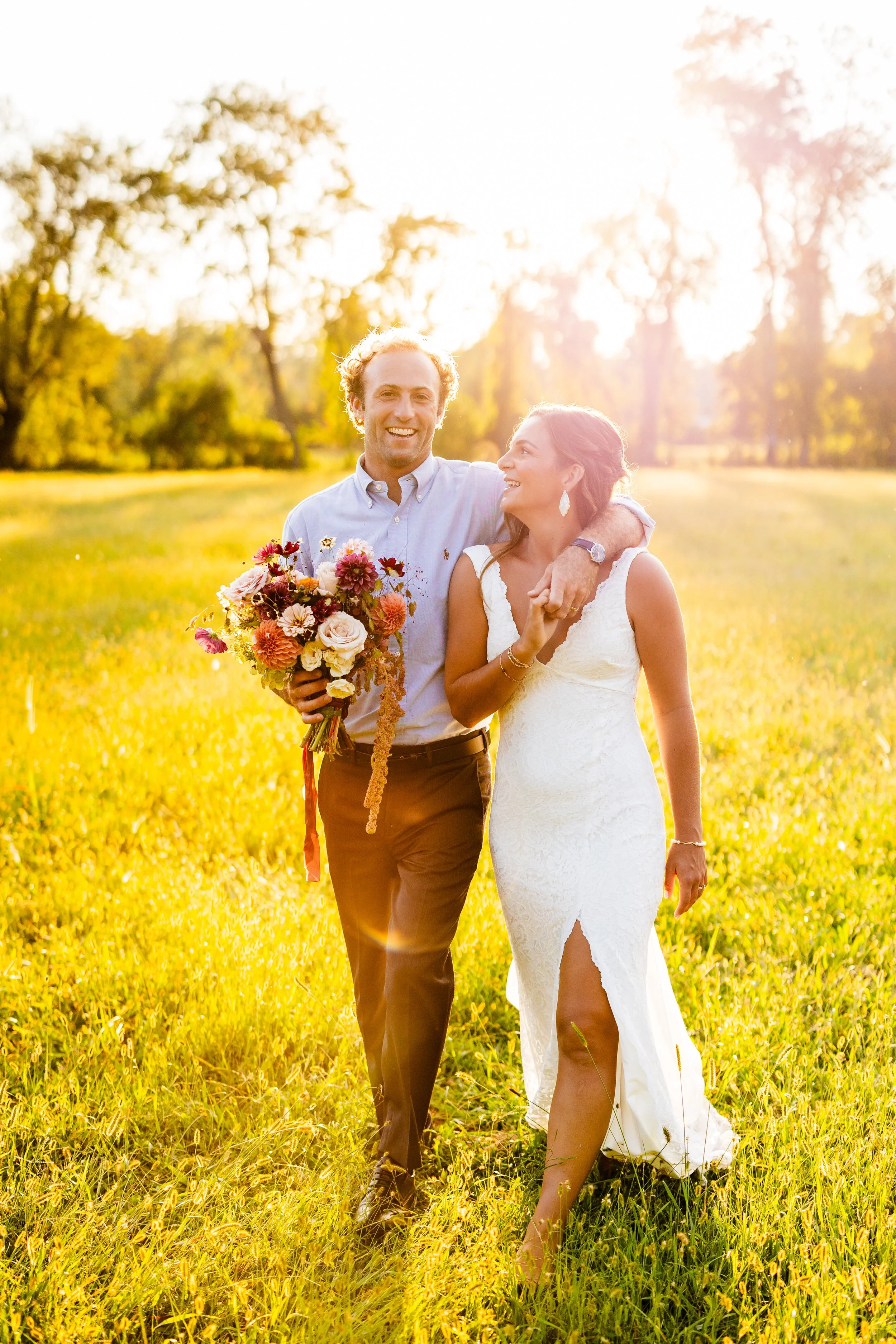 A smiling couple, a man and woman, walking arm-in-arm through a sunlit grassy field during sunset or golden hour, with the man holding a vibrant bouquet of flowers.