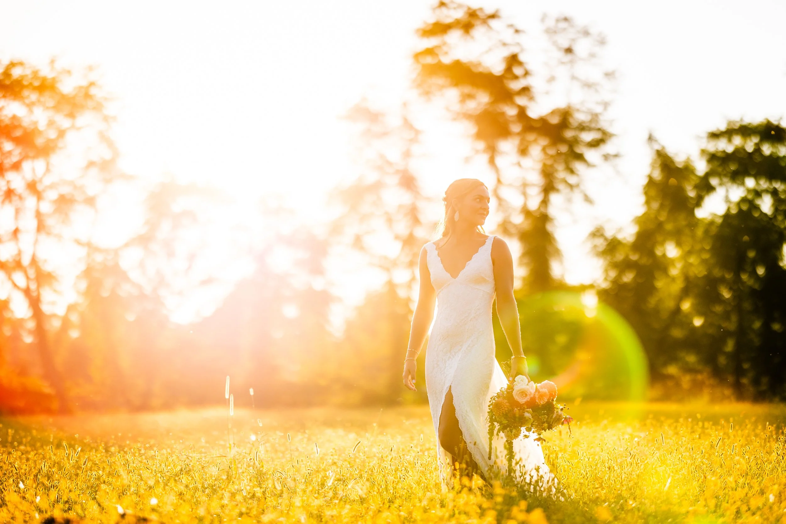 A woman in a white wedding dress walking through a grassy field at sunset, holding a bouquet of flowers, with trees in the background.