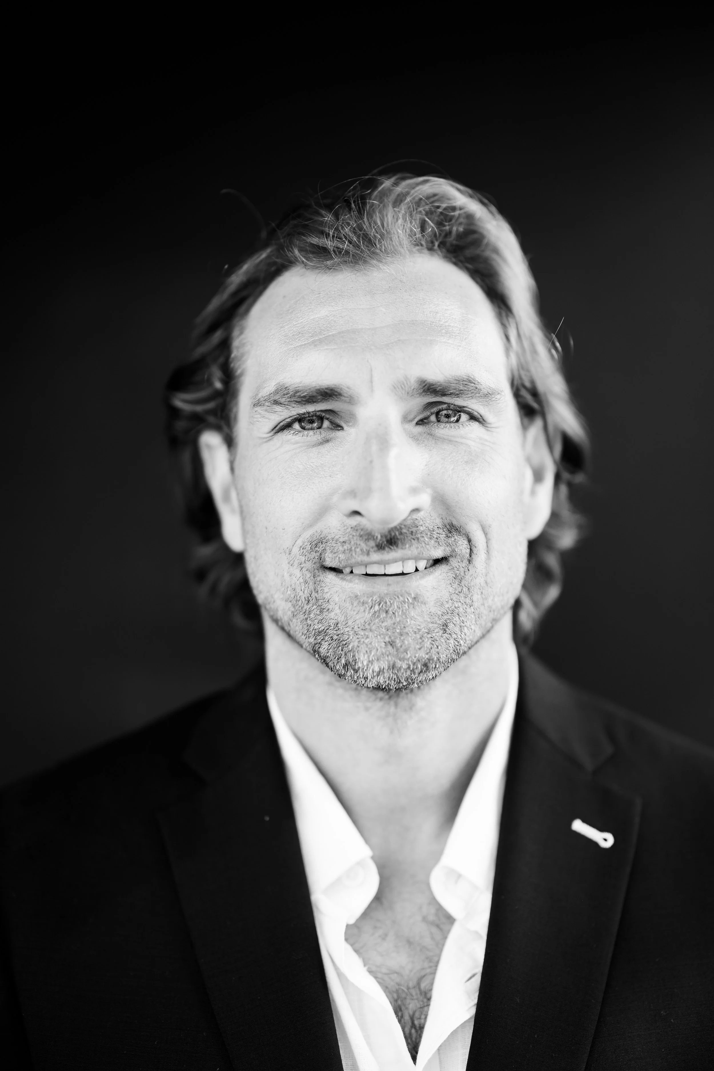 Black and white portrait of a smiling man with light-colored eyes, medium-length wavy hair, wearing a dark blazer and an open-collared shirt.