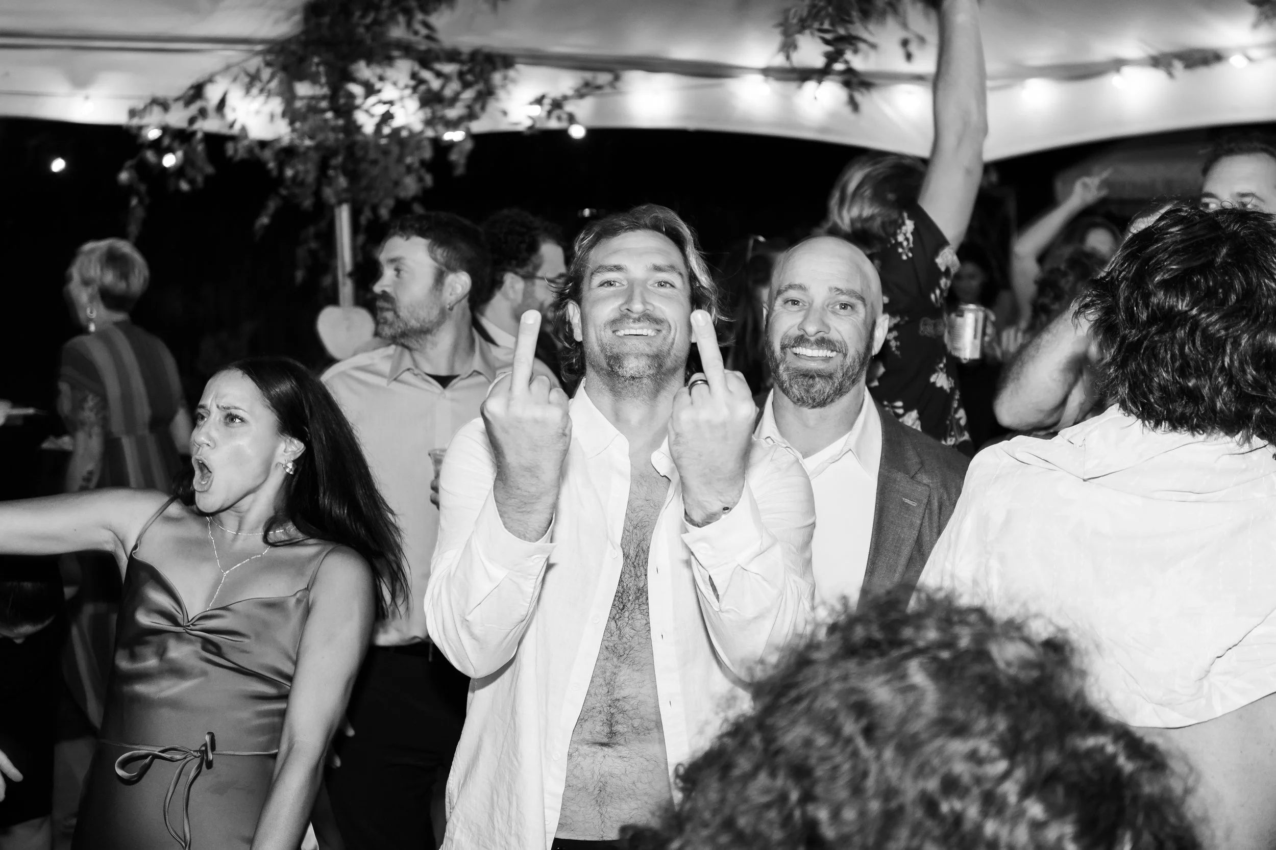Group of people at a party, with two men in the center smiling and showing middle fingers, and a woman on the left making a loud expression, all under a decorated tent with string lights.