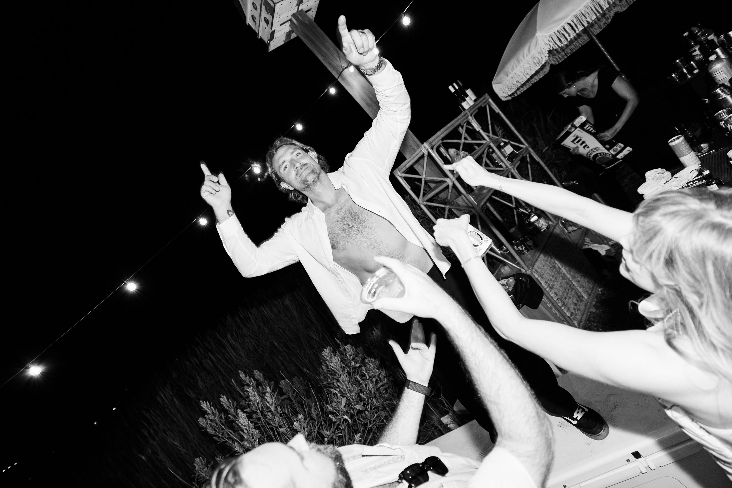 A man with a shirt open and chest hair visible, holding his arm up with both index fingers pointing upward, standing among people reaching out to him at an outdoor night event with string lights overhead.