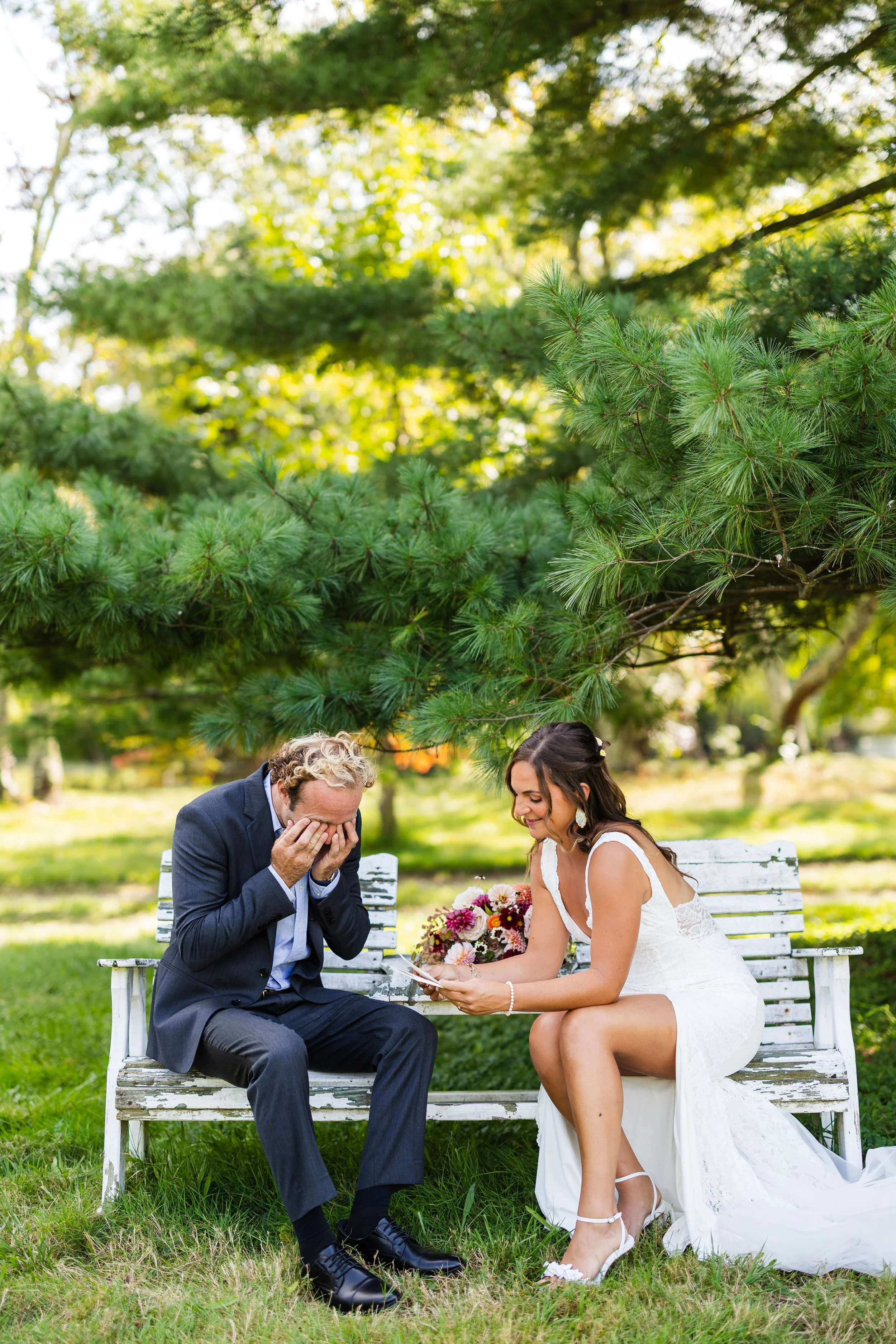 A bride in a white wedding dress and a man in a suit sit on a white wooden bench outdoors under green trees, sharing a joyful moment during a wedding.