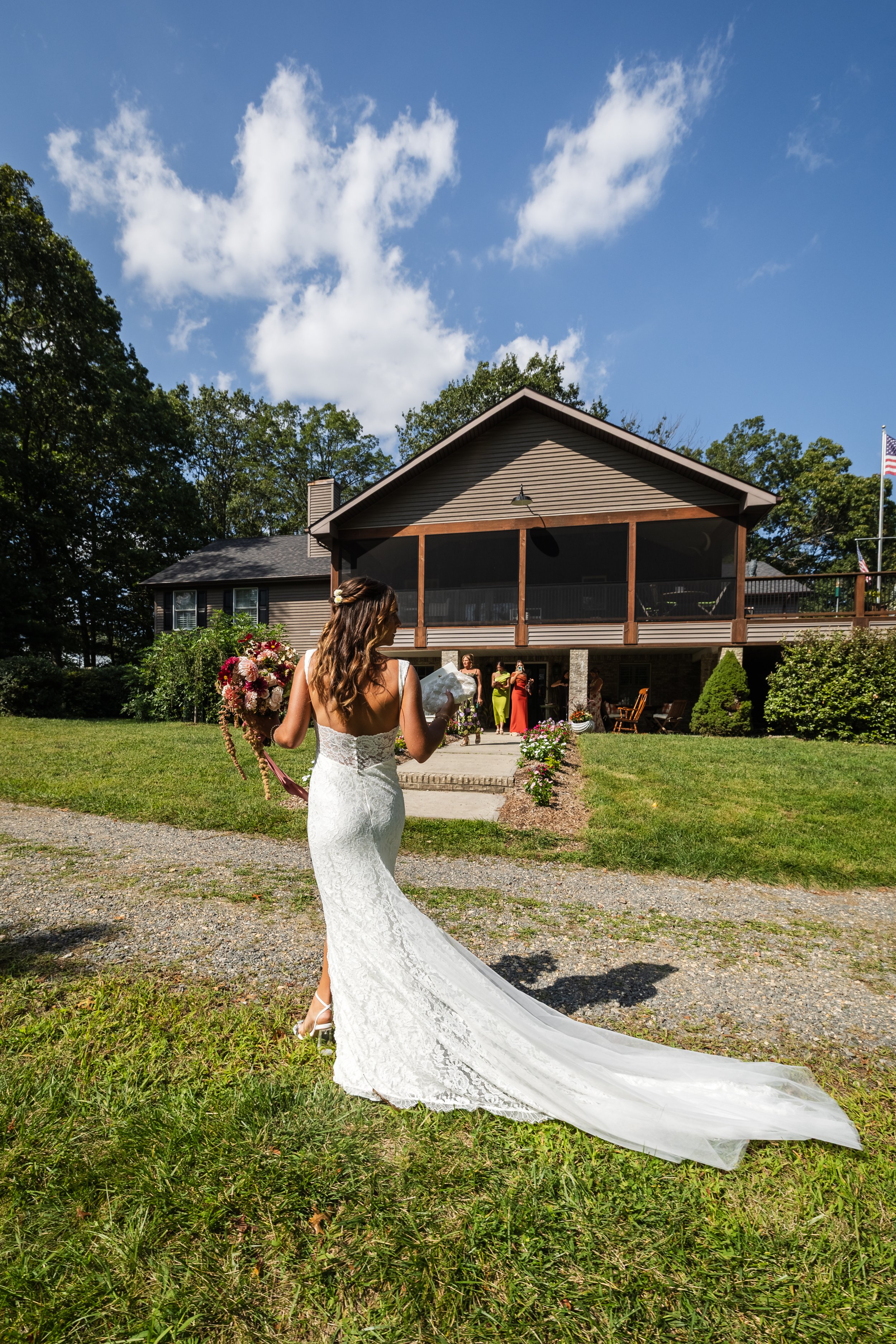 Bride in a white lace wedding dress walking towards a house with guests in the background on a sunny day.