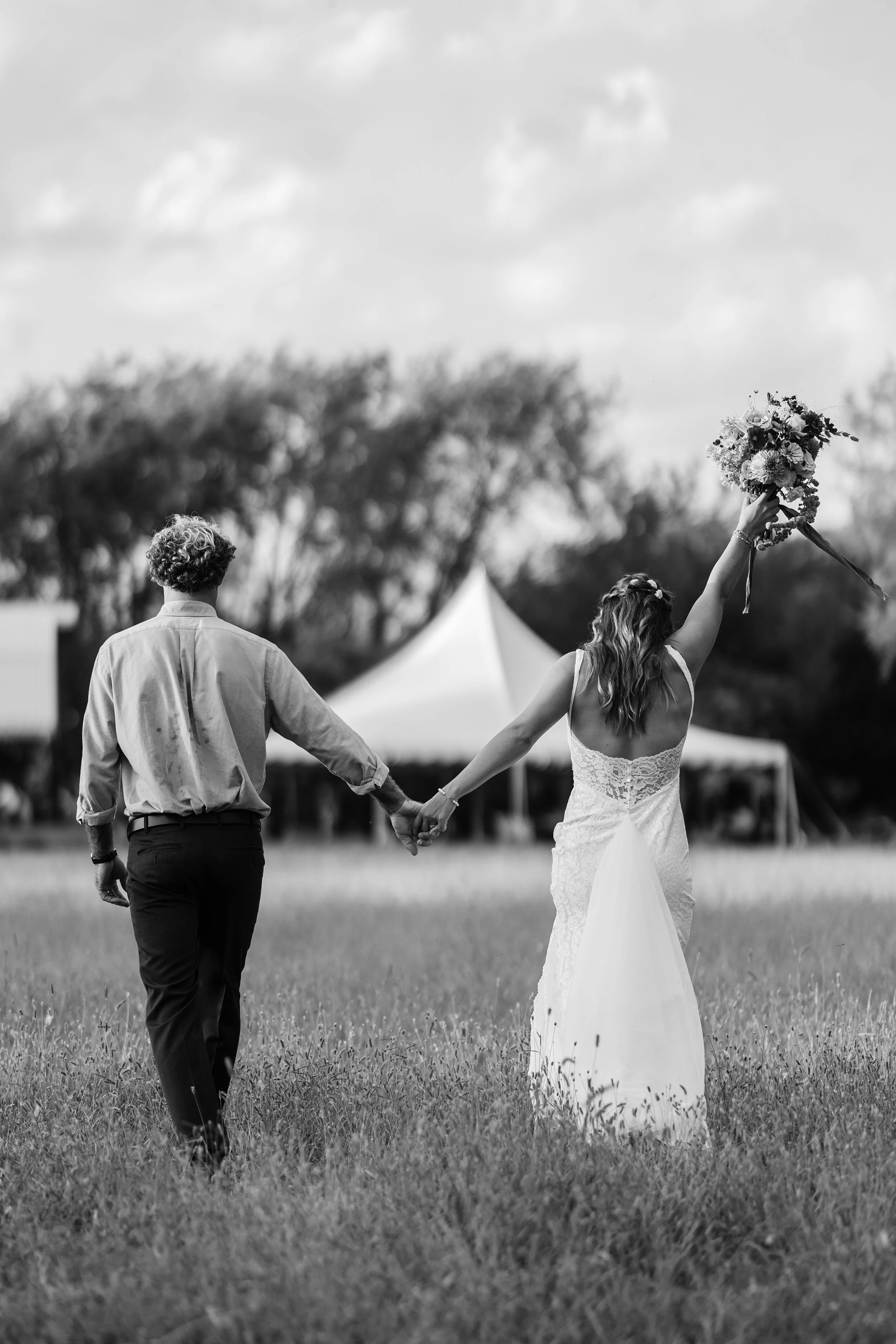 Black and white photo of a newlywed couple walking hand in hand on a grassy field, the bride raising a bouquet in the air.