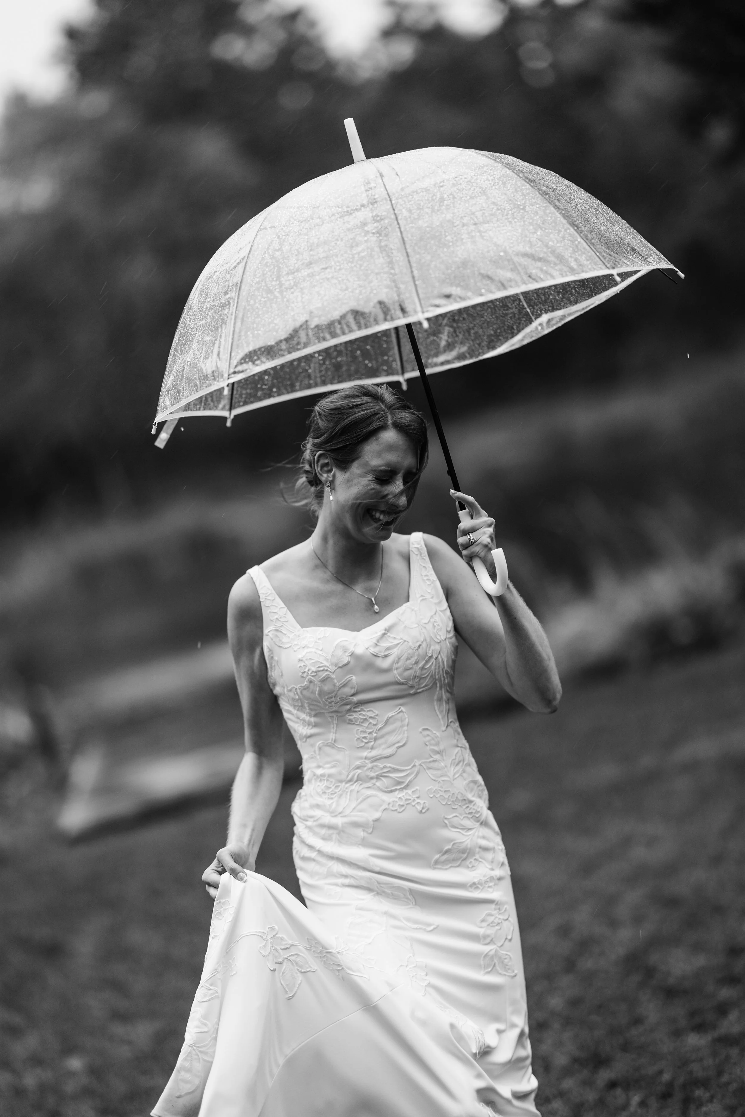 A woman in a wedding dress holding an umbrella and smiling in the rain outdoors.
