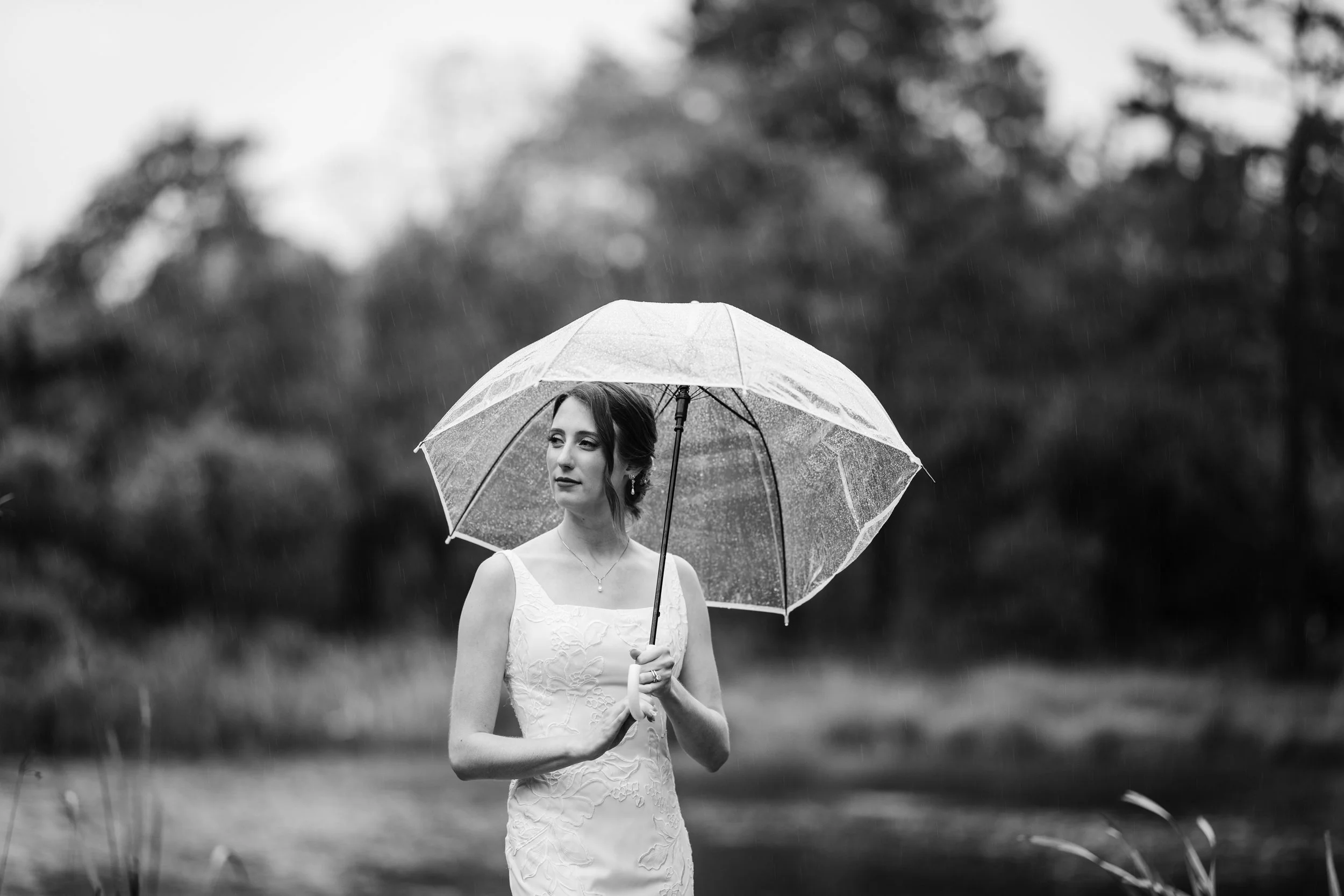 A woman in a wedding dress holding a transparent umbrella outdoors in the rain, with trees in the background.