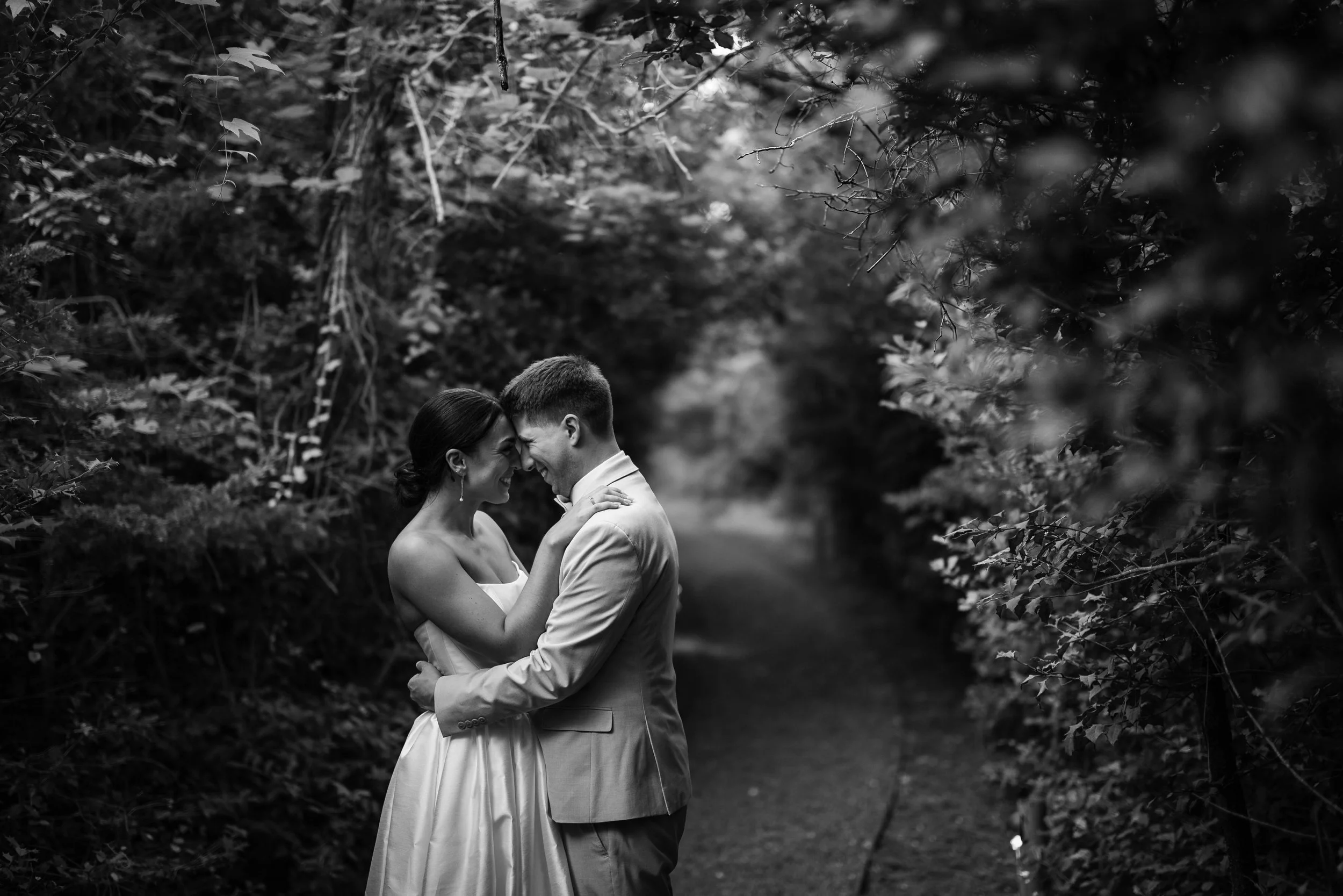 A black and white photo of a couple on their wedding day, standing close together with foreheads touching, smiling, and holding each other in a natural, wooded outdoor setting.