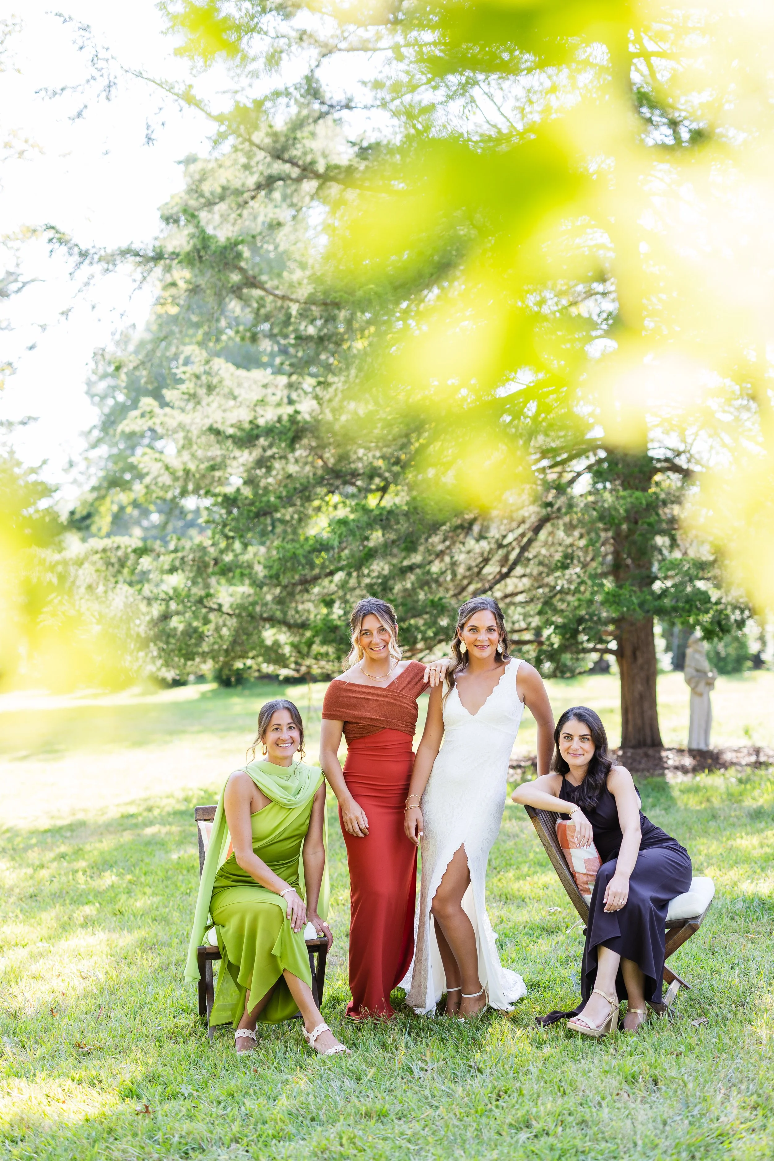Four women in elegant dresses outdoors, one standing and three seated on chairs, under a large tree on a sunny day.