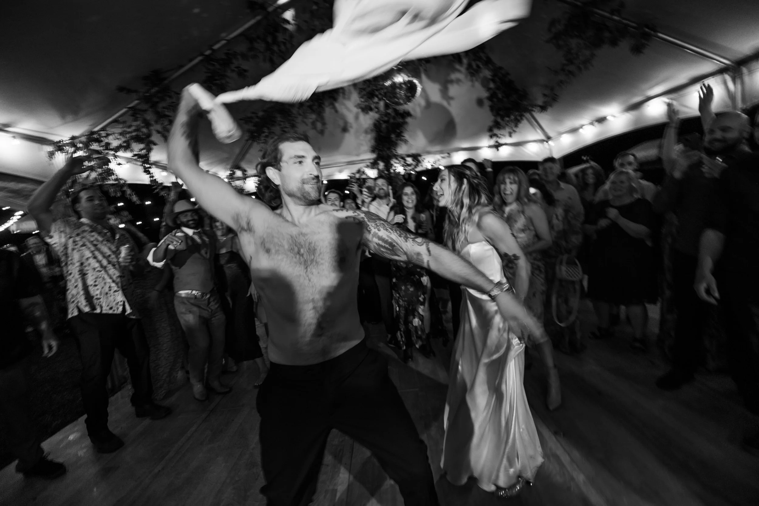 A black and white photo of a shirtless man dancing with a flowing cloth at a lively party, surrounded by smiling guests under a decorated tent ceiling.