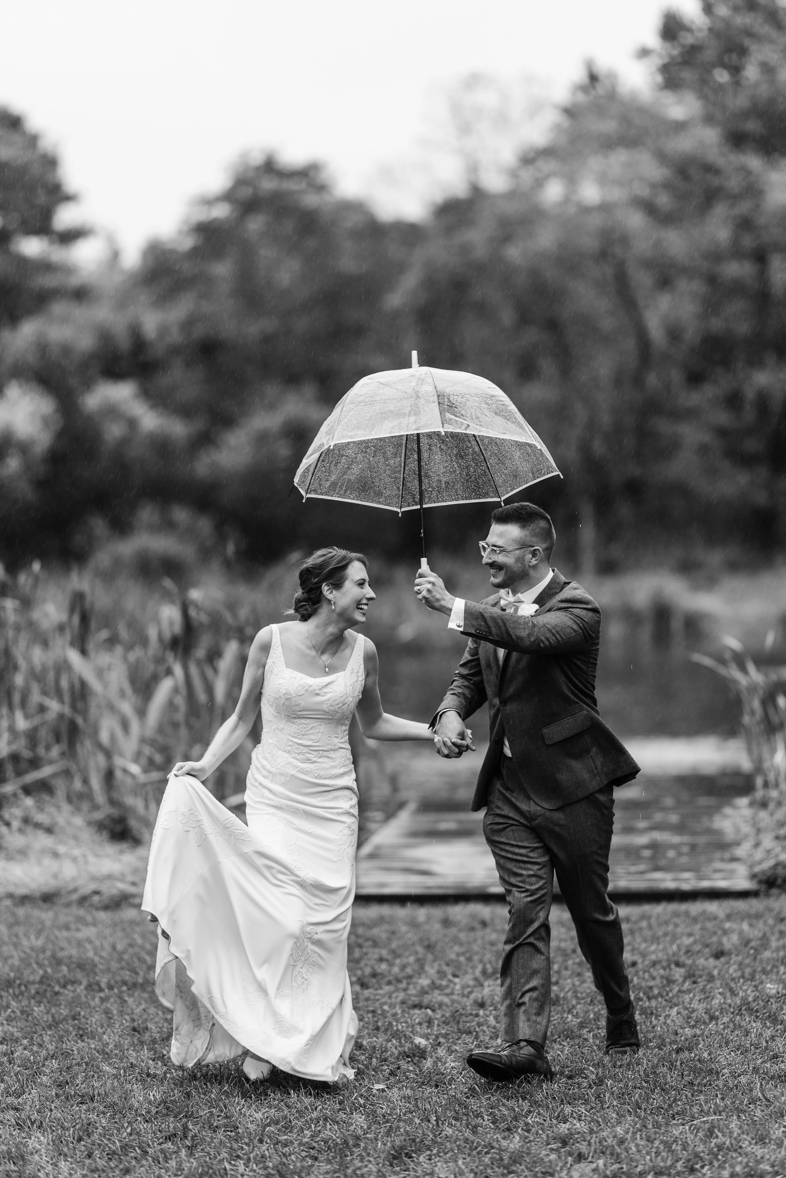 A happy couple in wedding attire running under an umbrella in the rain outdoors, smiling and holding hands.