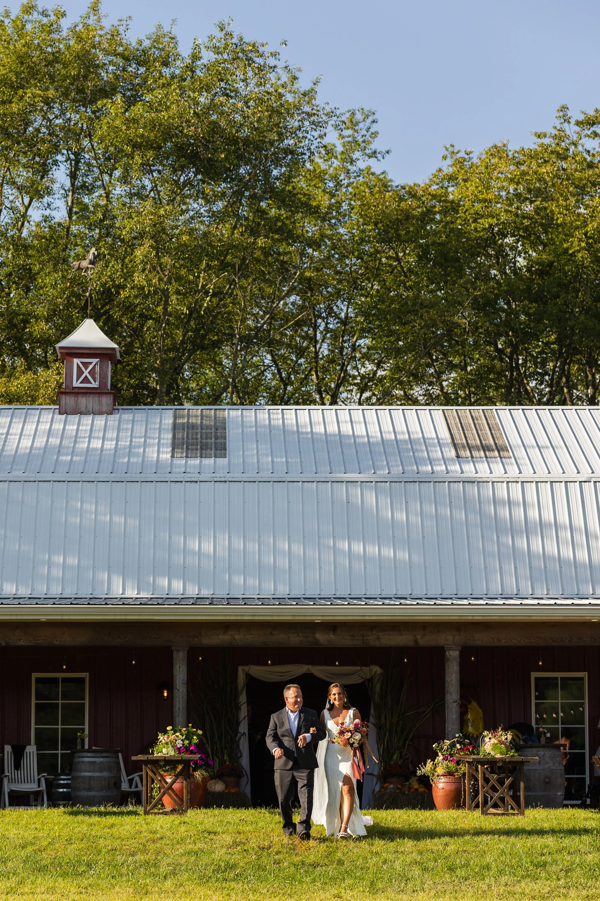 A bride walking down the aisle with her father in front of a barn decorated with flowers on a sunny day.