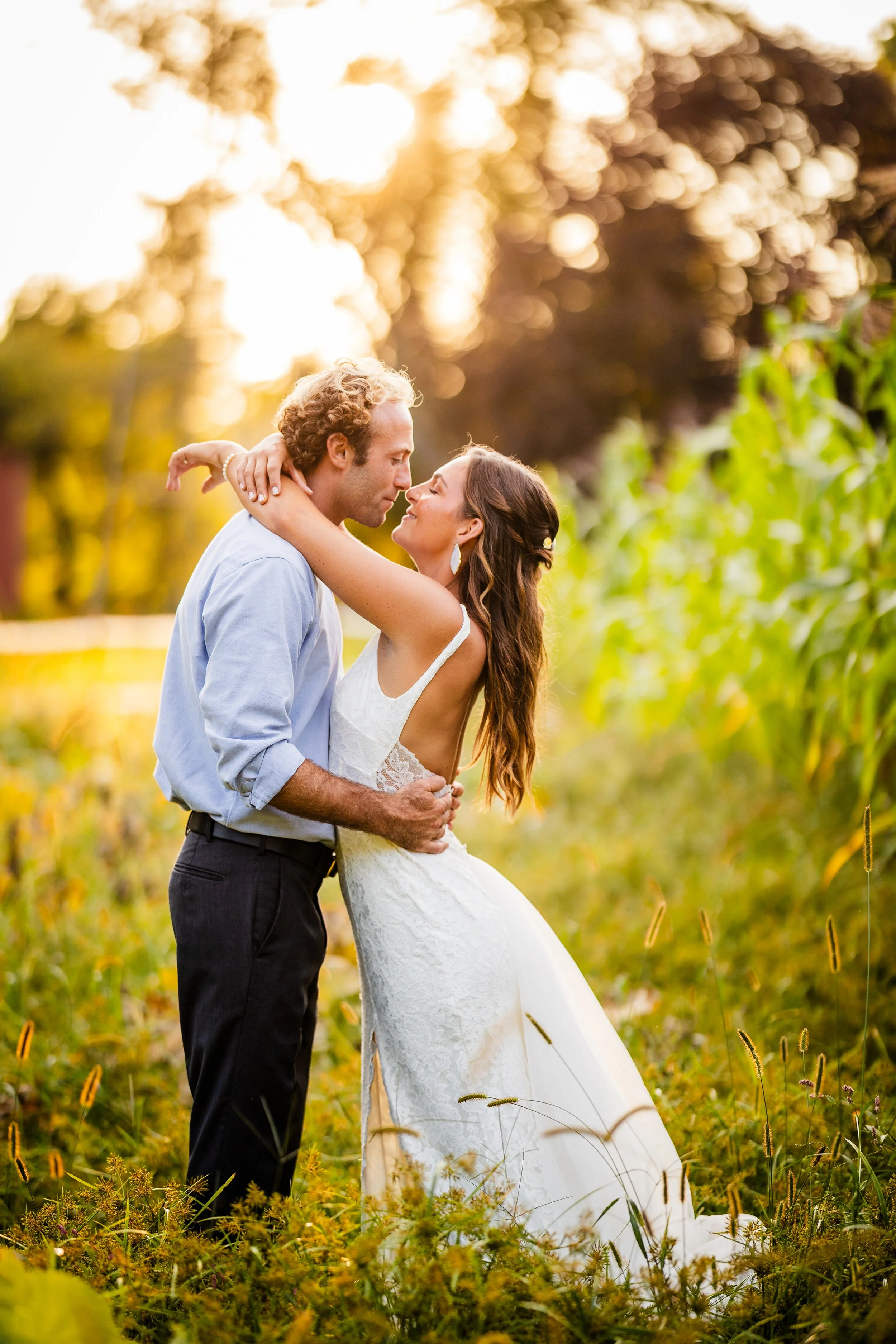 A couple dressed in wedding attire stands in a field during sunset, embracing each other closely, with trees and warm sunlight in the background.