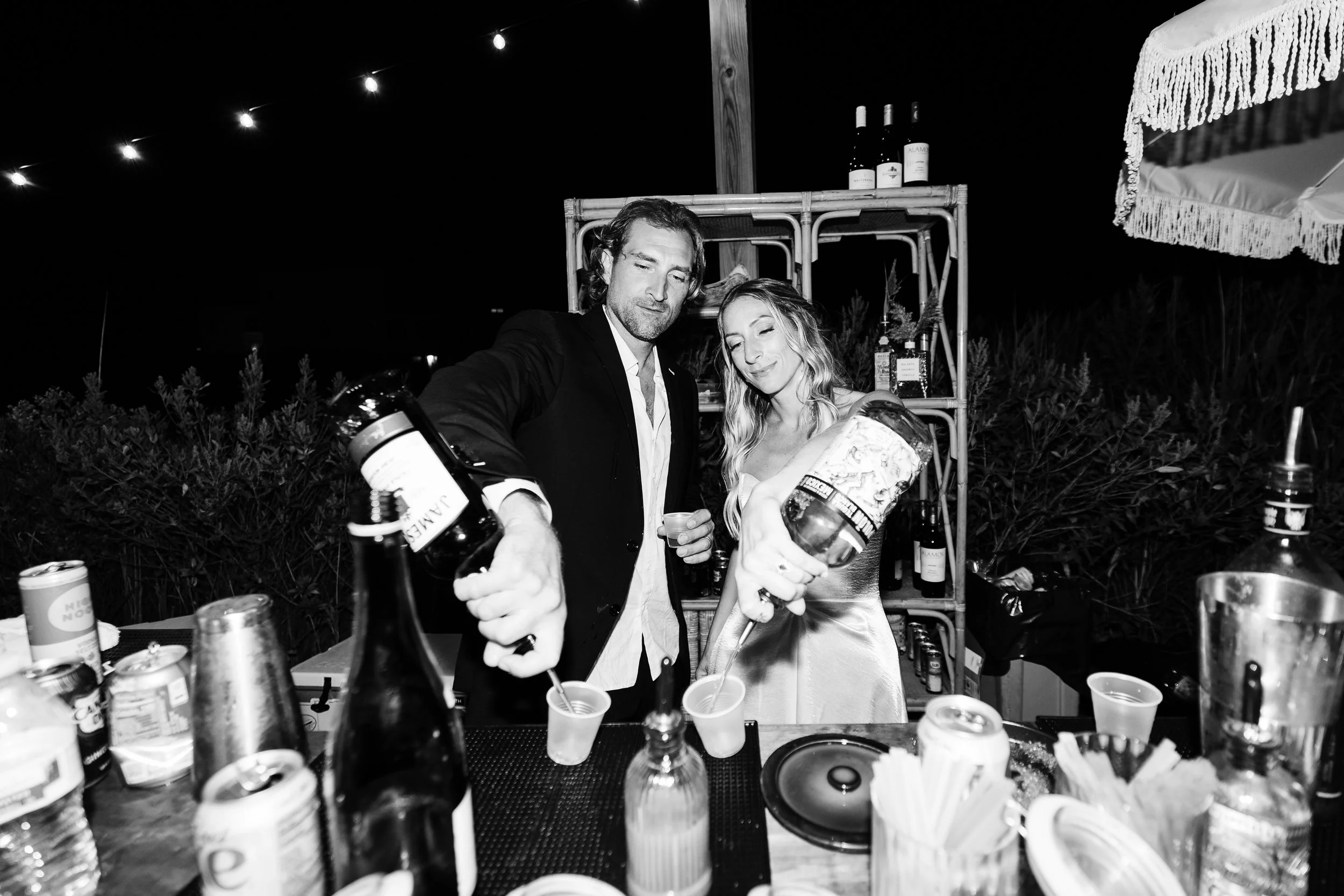 A man and woman pouring drinks into cups at an outdoor party at night, with bottles and snacks on the table, under string lights.