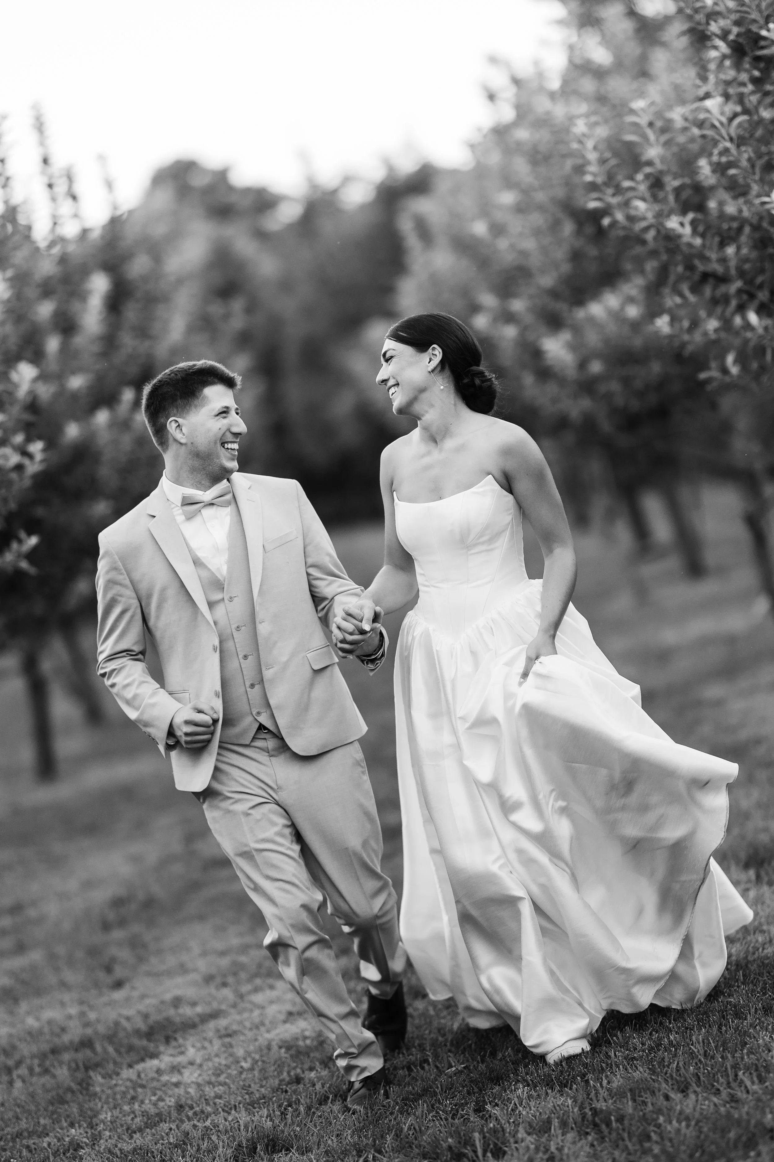A groom and bride holding hands and laughing outdoors in a park or garden setting, wearing a suit and wedding dress.