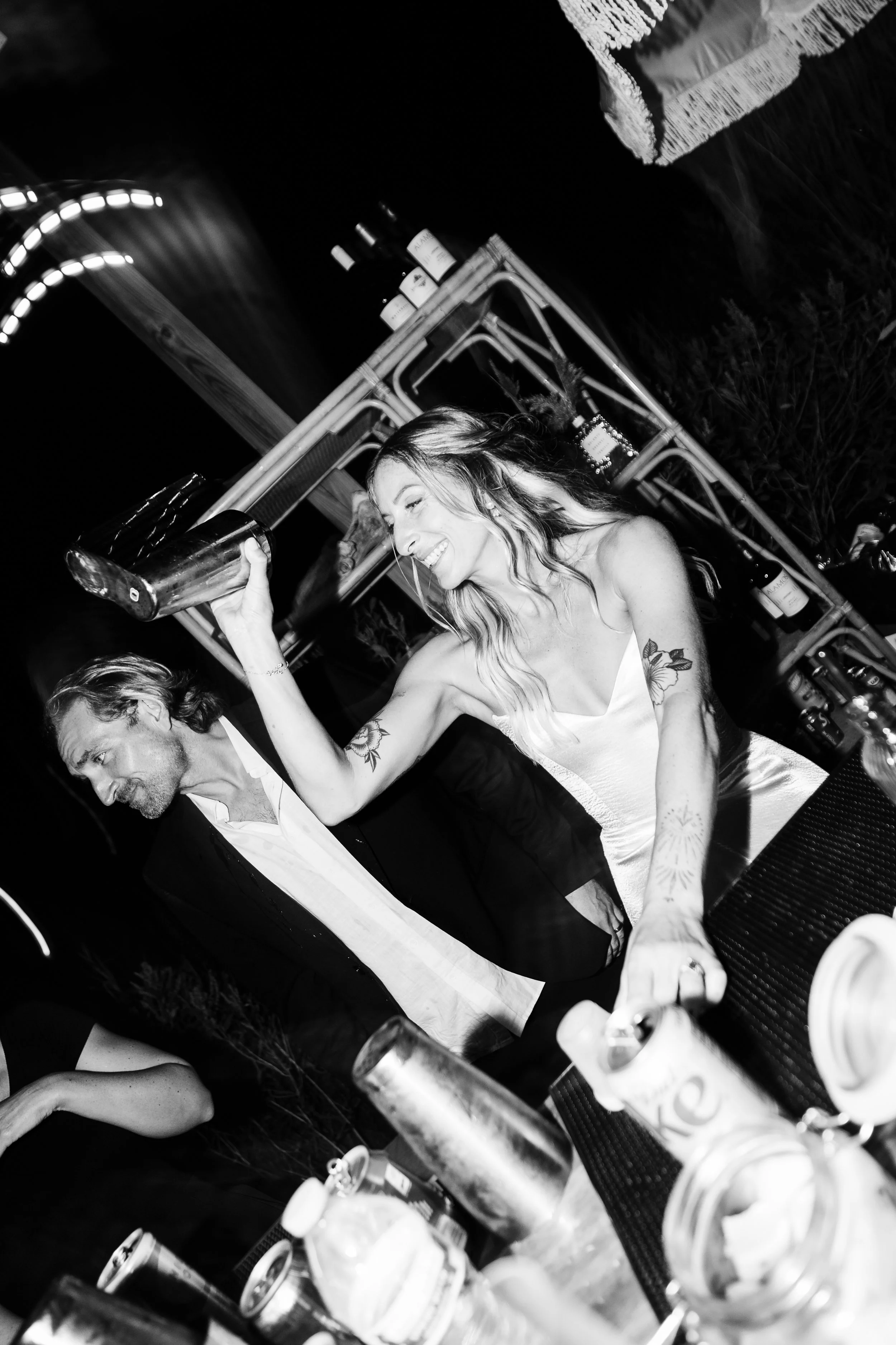A woman smiling and dancing with a drink in her hand at a party with bottles, cans, and glasses on a table.