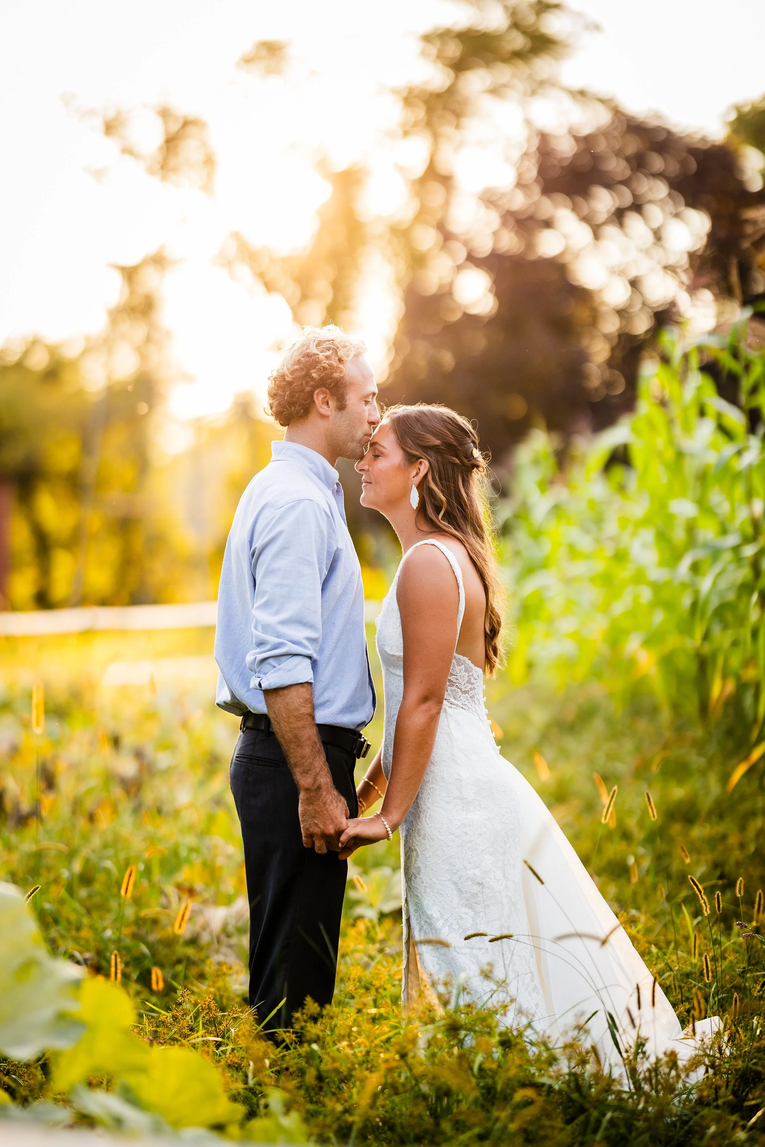 A bride and groom standing close in a field at sunset, holding hands and touching foreheads.