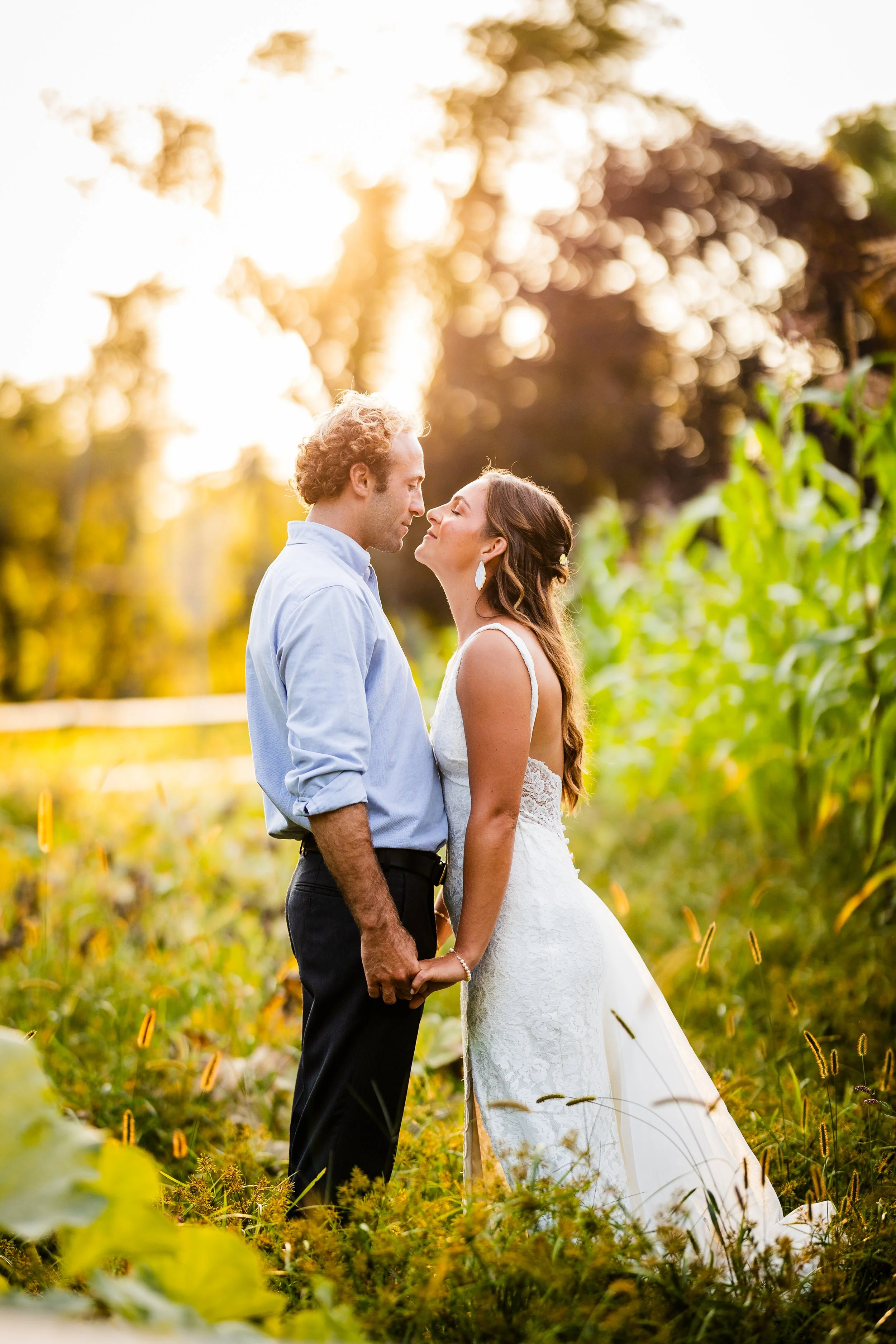 A bride and groom standing close together outdoors in a field during sunset, holding hands and facing each other affectionately.