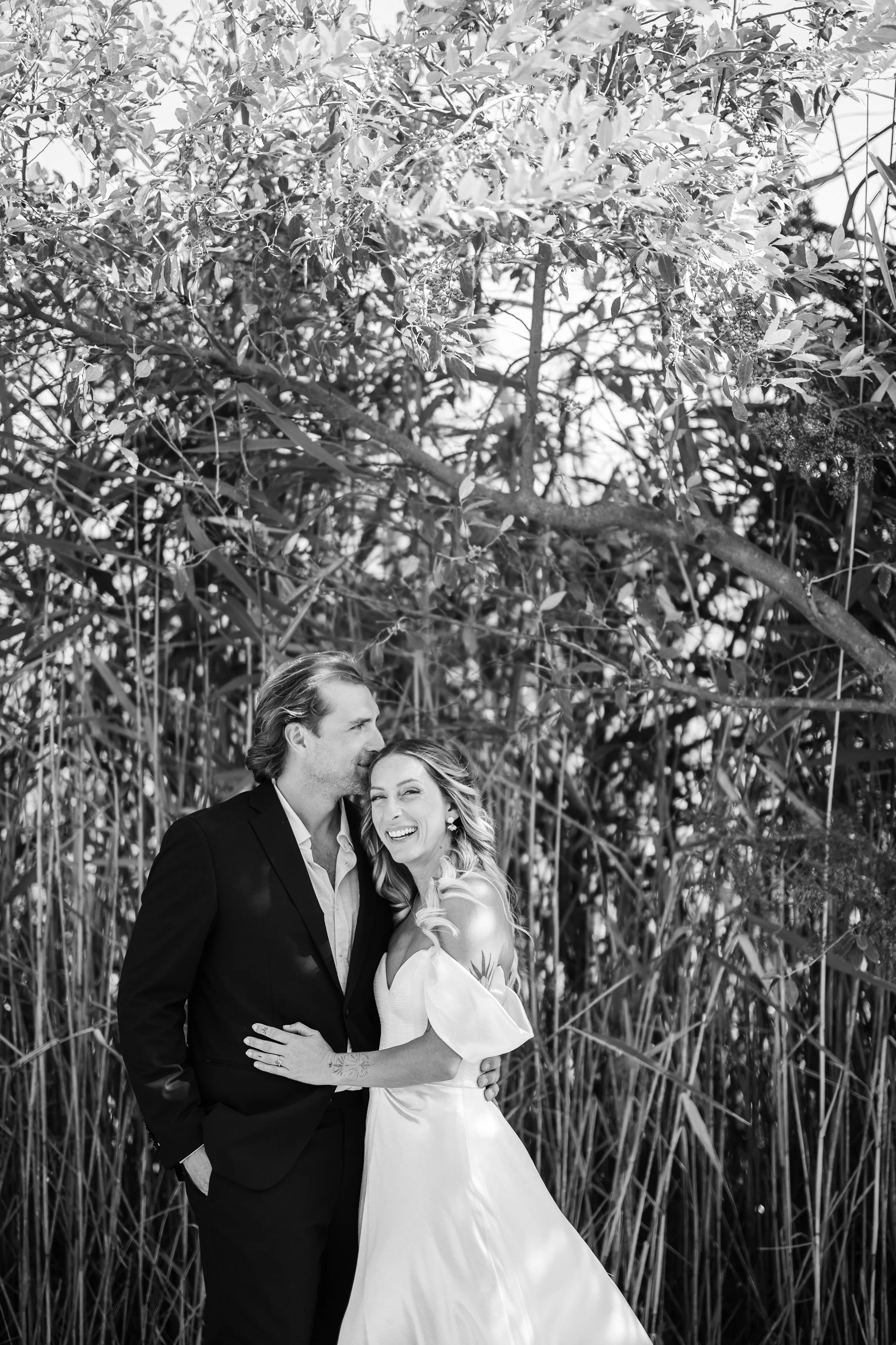 A black and white photo of a couple, dressed in formal attire, standing close together outdoors among tall grass and trees, smiling and embracing each other.