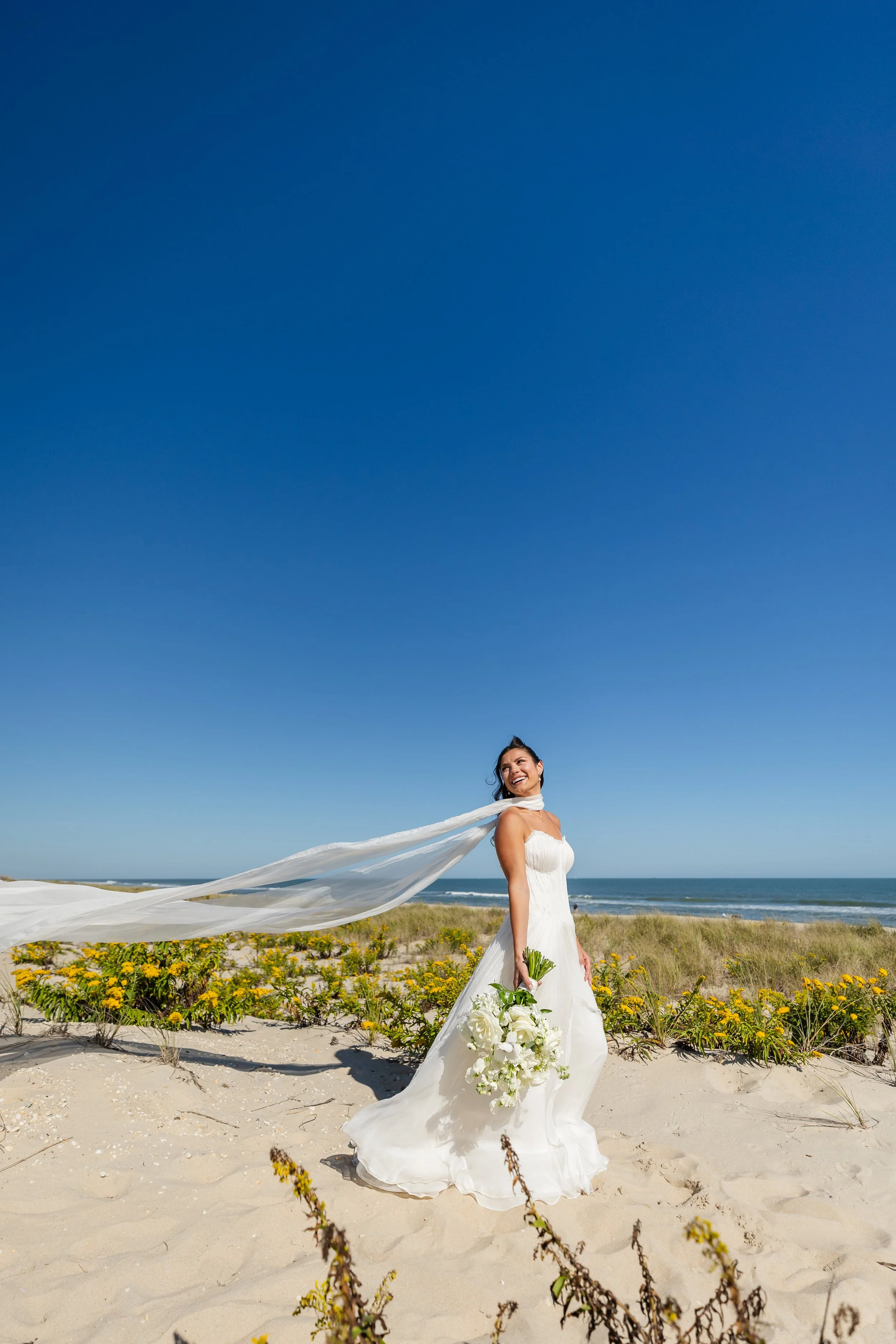 A bride in a white wedding dress holding a bouquet of white flowers, smiling and standing on the beach.