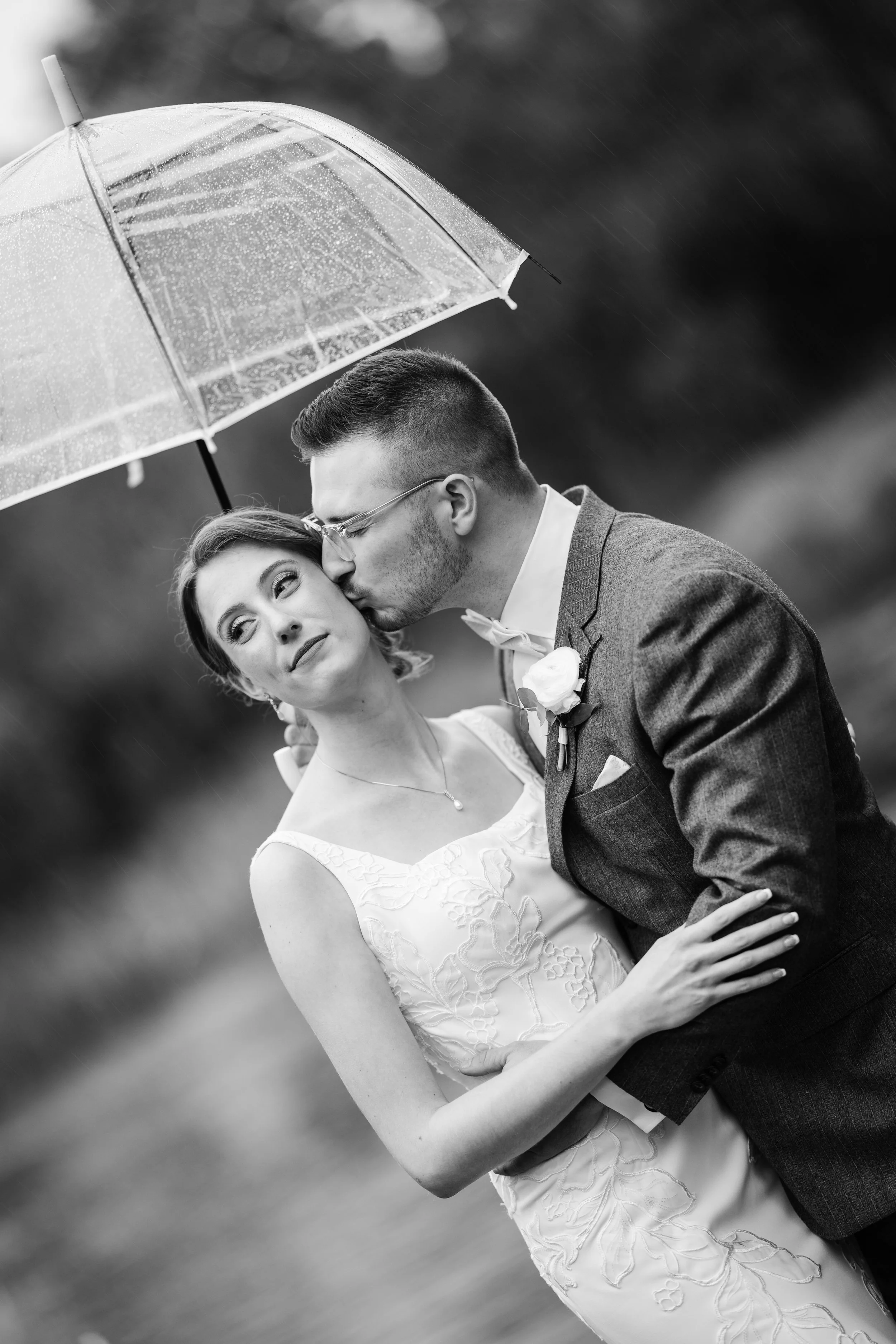 A black and white photograph of a newlywed couple under a clear umbrella during rain, with the groom kissing the bride on the cheek as she looks into the distance.