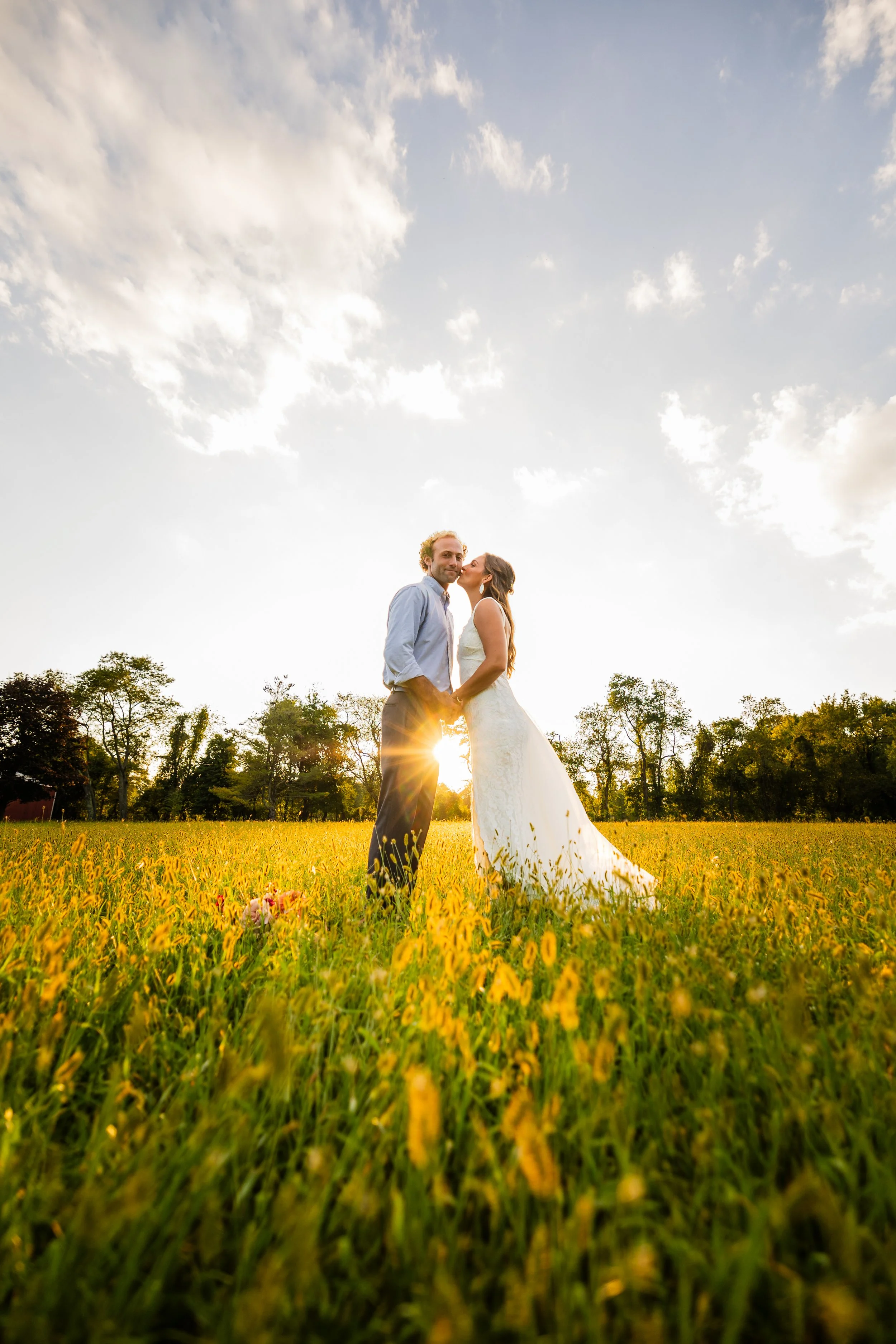 Couple in wedding attire standing in a yellow flowered field at sunset, facing each other and holding hands, with sun shining behind them and trees in the background.