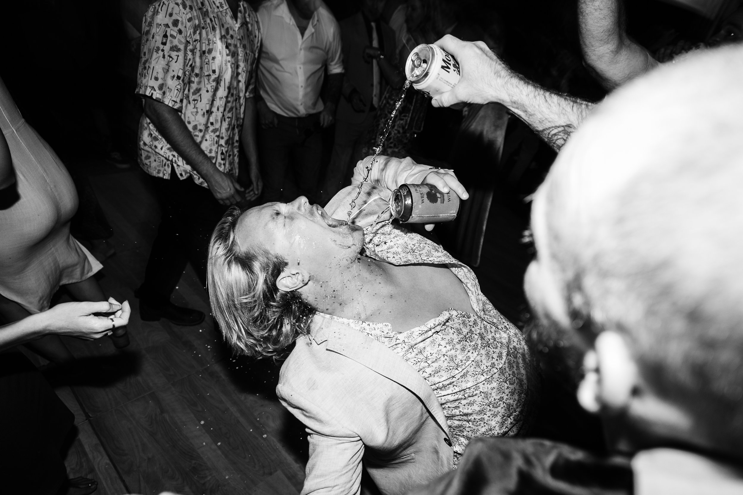 A woman is lying on her back with her head tilted back, while a man pours a drink into her mouth during a party or celebration.