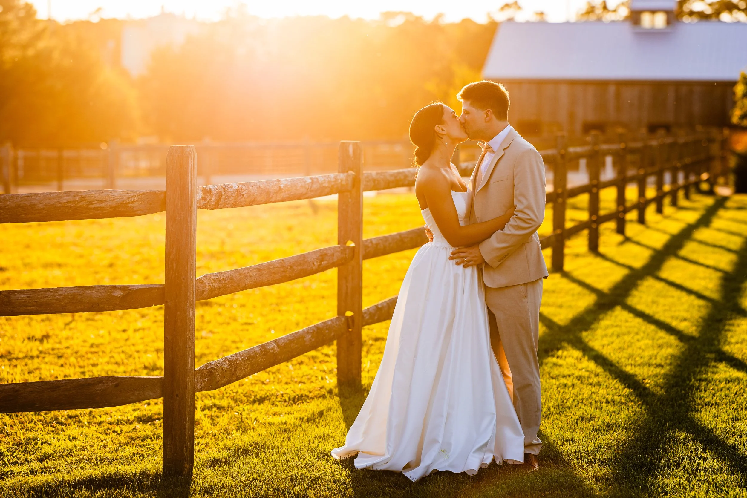 A bride and groom kissing outdoors during sunset, with a wooden fence and a barn in the background.