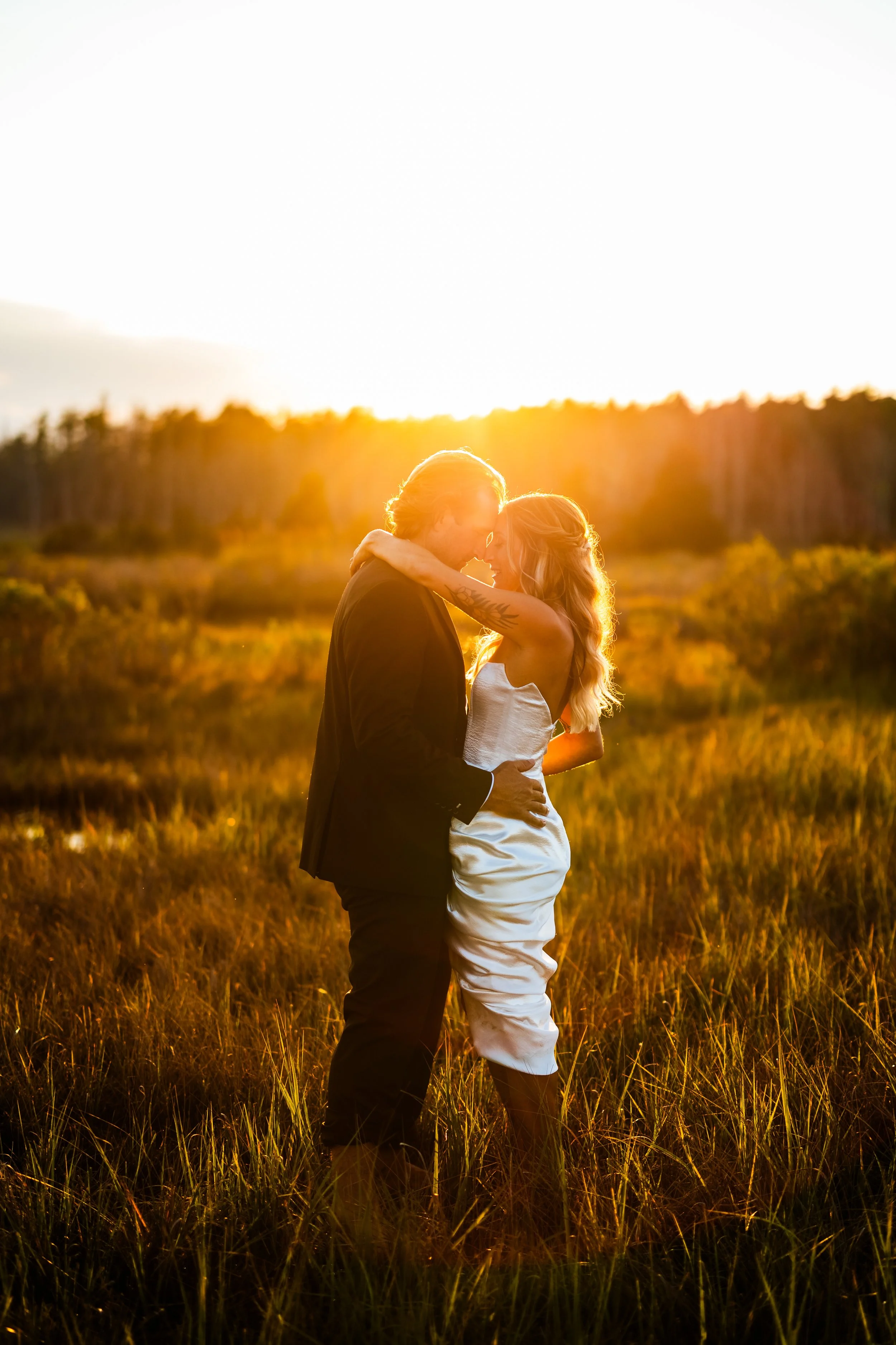 A couple embracing in a field at sunset, with their foreheads touching and the sun setting behind them.