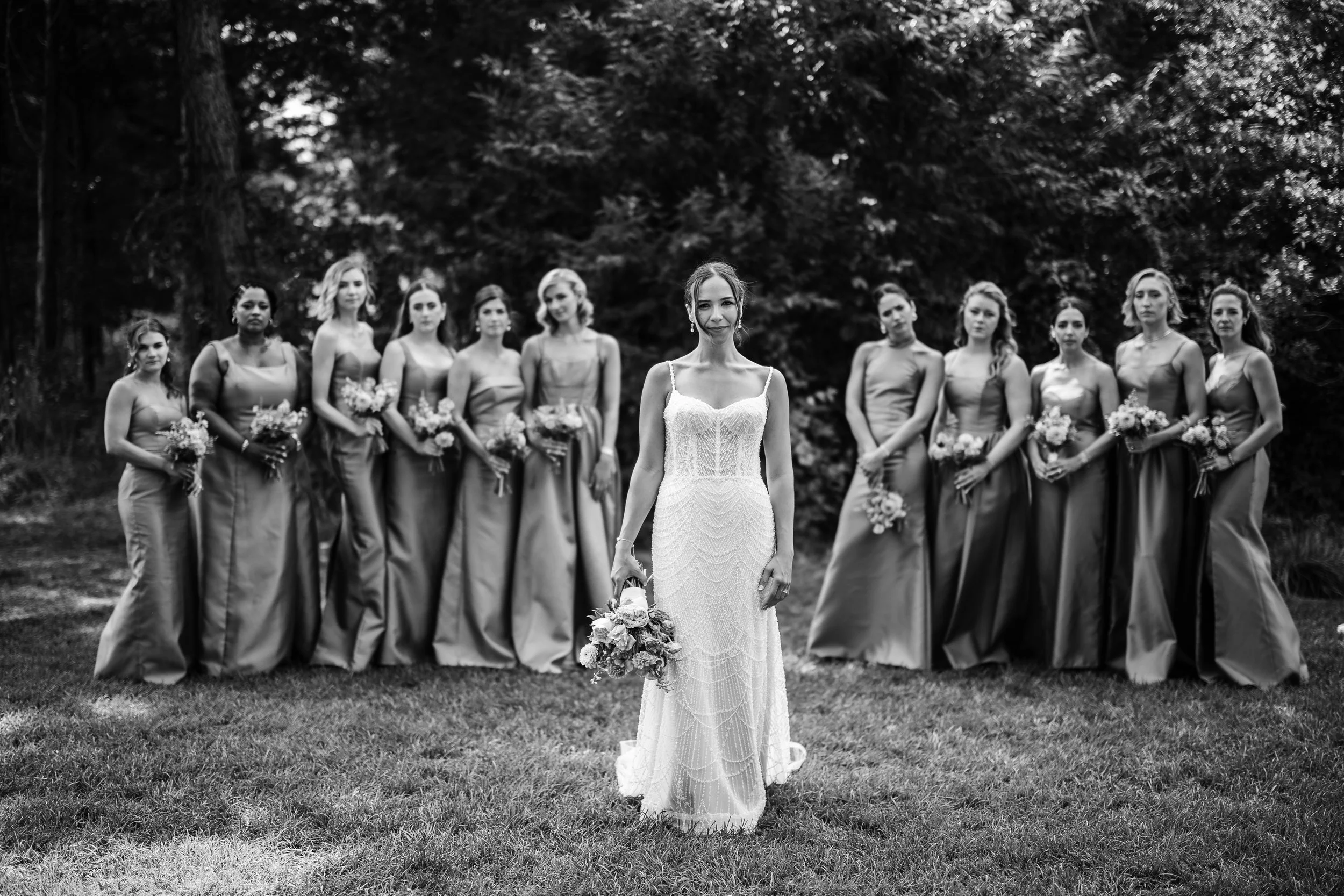 Black and white photo of a bride in a wedding dress holding a bouquet standing in front of twelve bridesmaids in long dresses, outdoors with trees in the background.