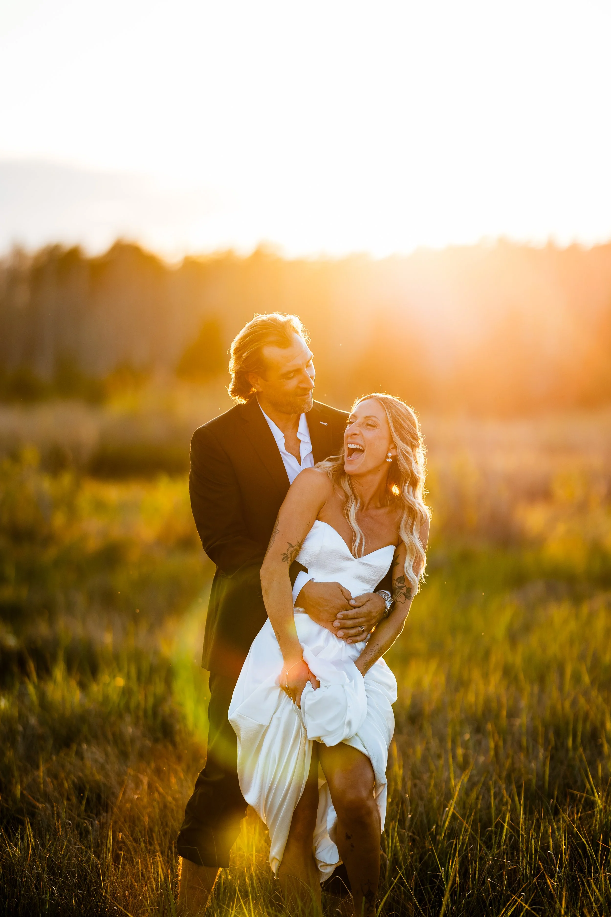 A couple dressed in wedding attire sharing a joyful moment outdoors at sunset. The woman is wearing a white dress and the man a dark suit, standing in a grassy field with trees in the background.
