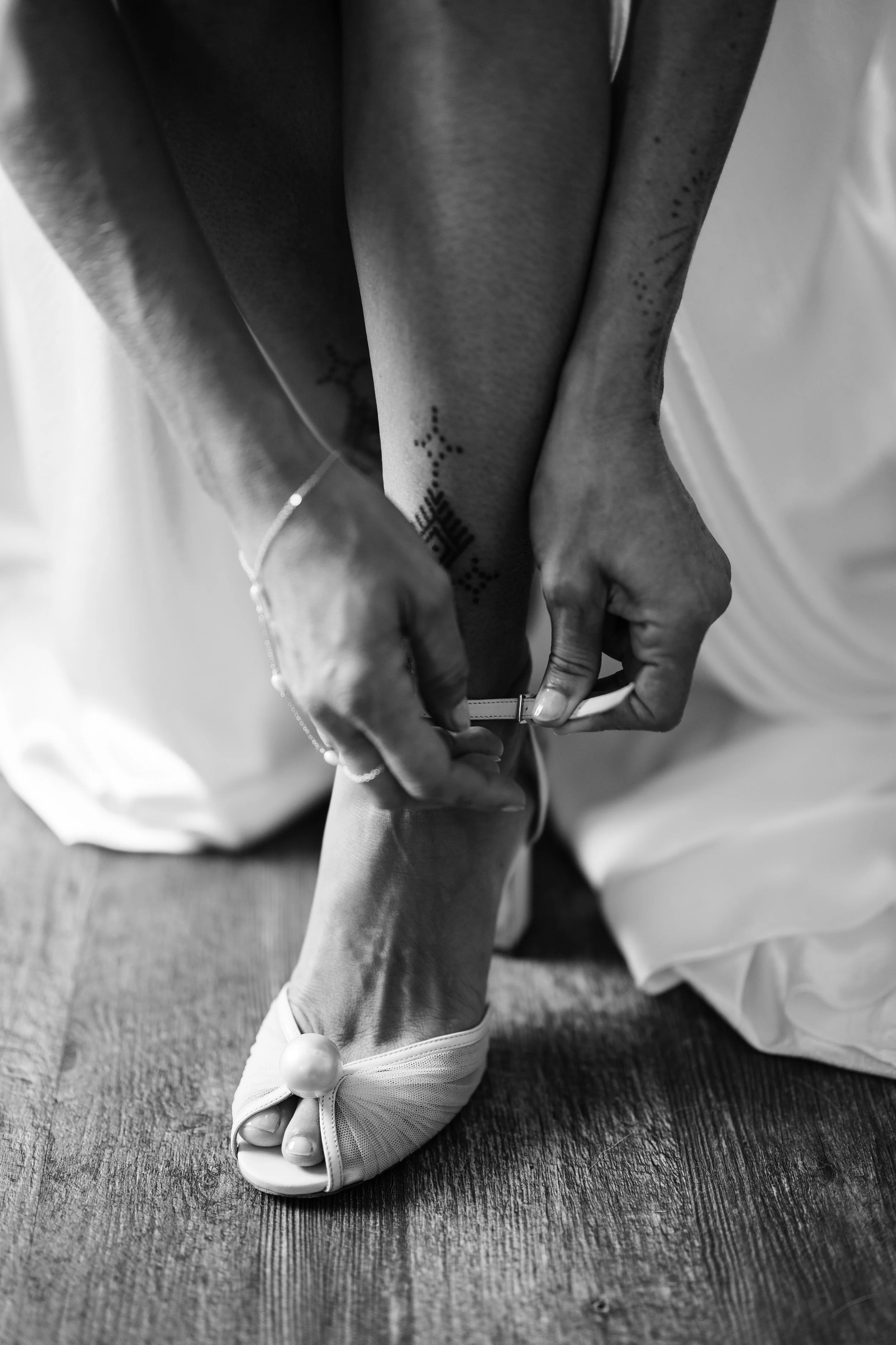 A person adjusts a garter on their leg, wearing a wedding shoe and a delicate bracelet, with tattoos visible on their arm.