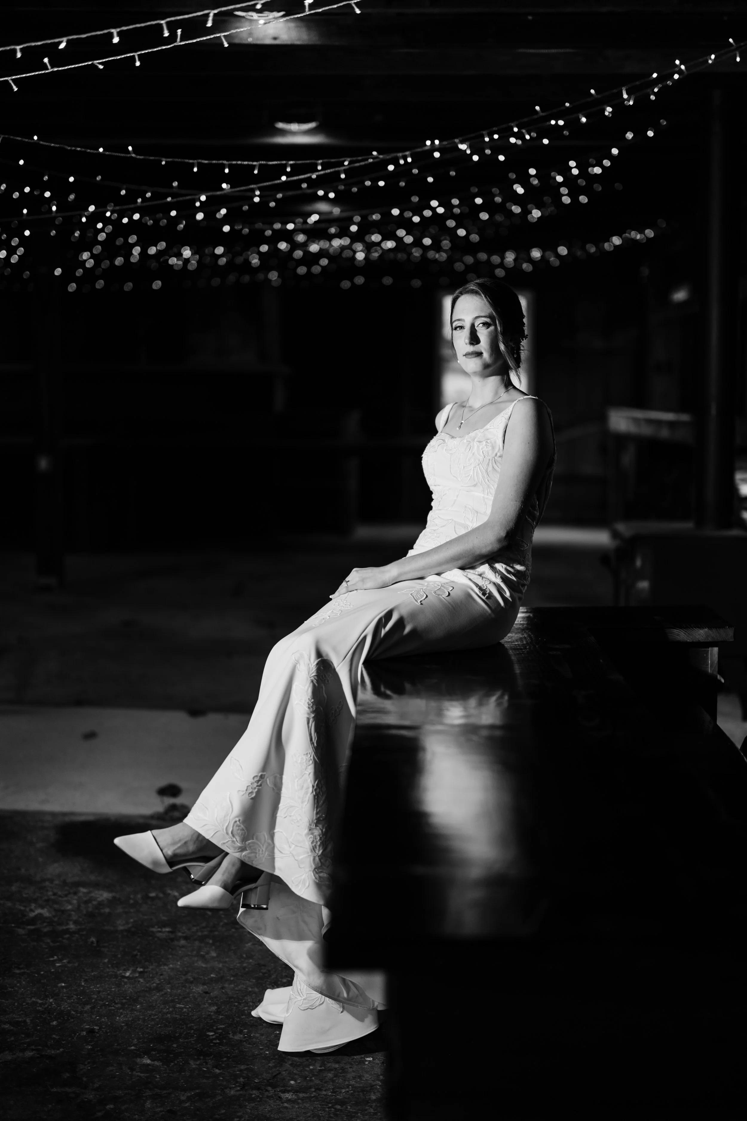 A woman in a wedding dress sitting on a wooden bench in a dimly lit space with string lights hanging from the ceiling.