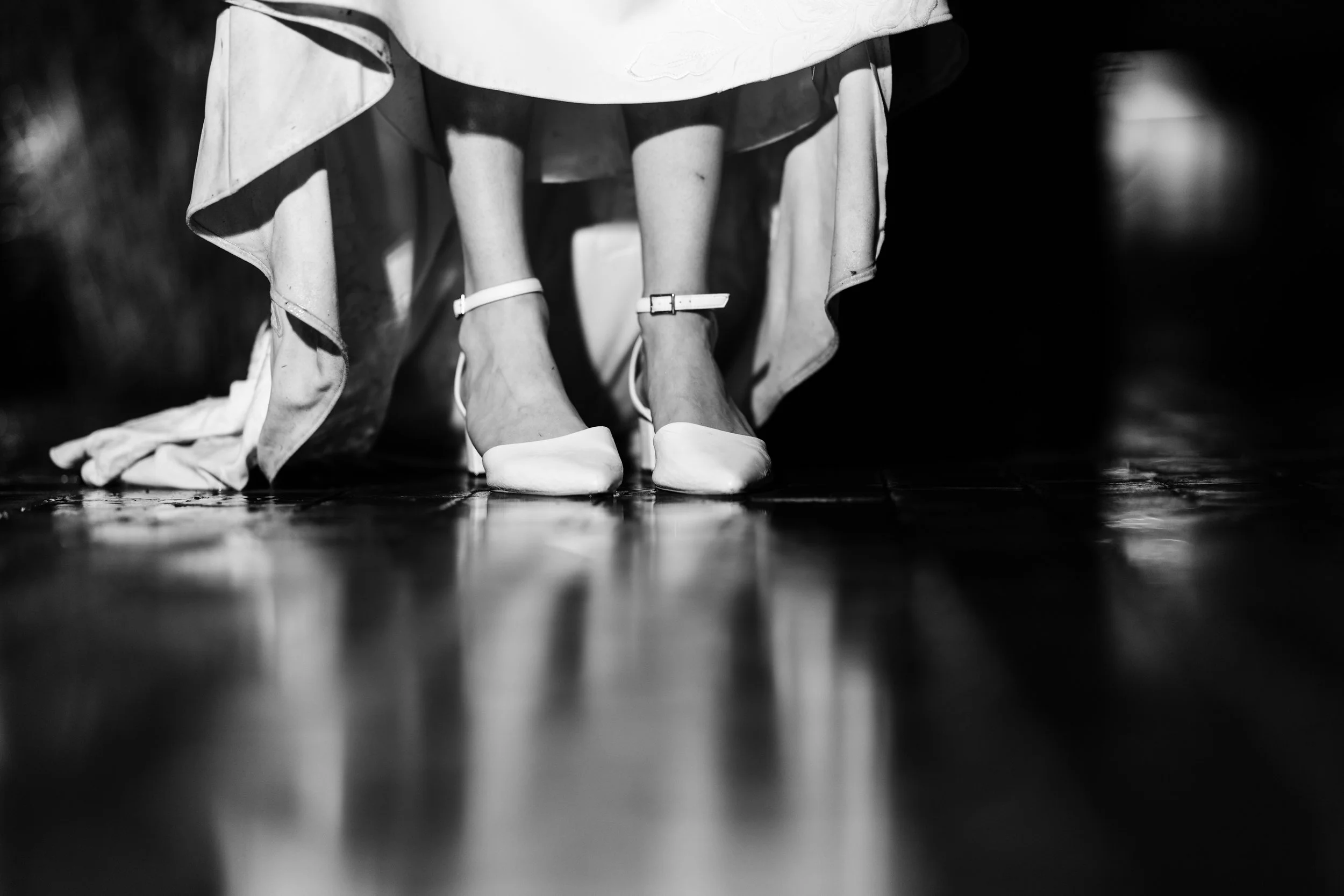 Black and white photo of a woman’s legs and feet, showing her wearing white high heels and a flowing skirt, with her standing on a shiny floor, possibly during a performance or event.