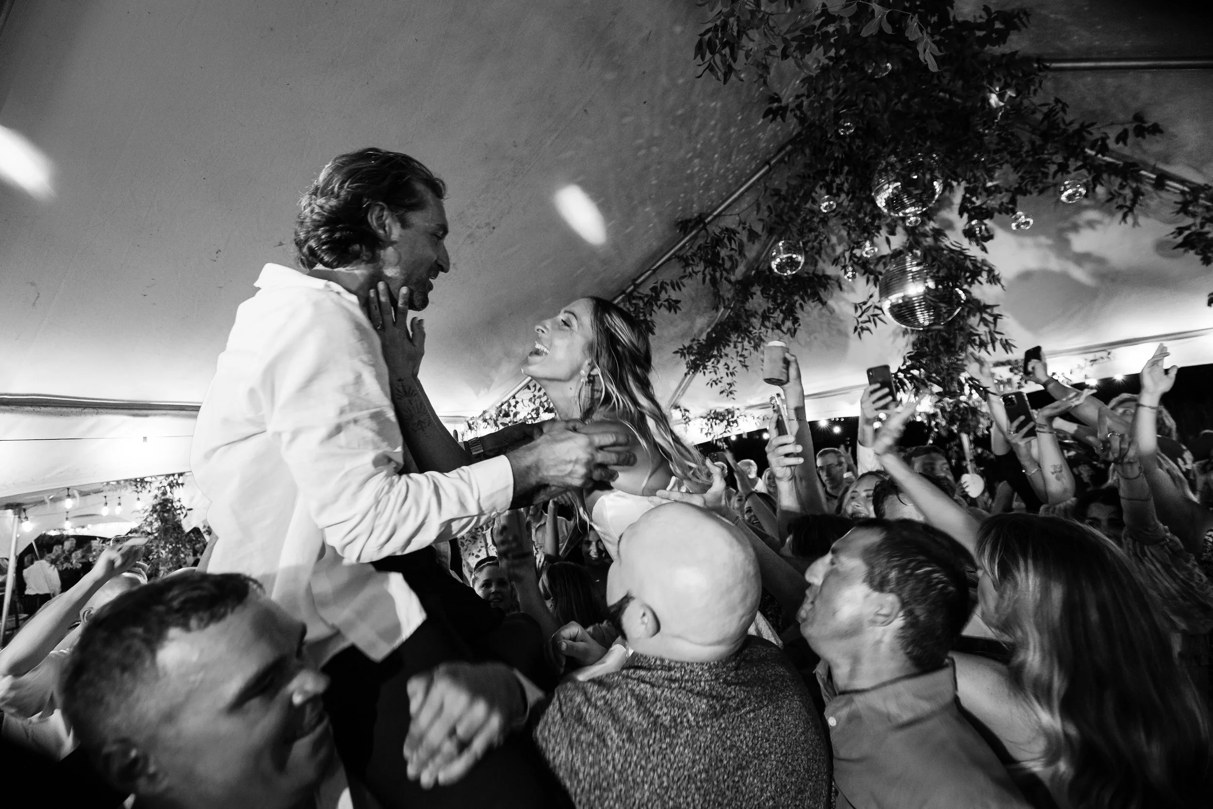 A joyful celebration at a party with a large crowd dancing and taking photos. A man and woman are on a platform, smiling and holding each other, with onlookers surrounding them.