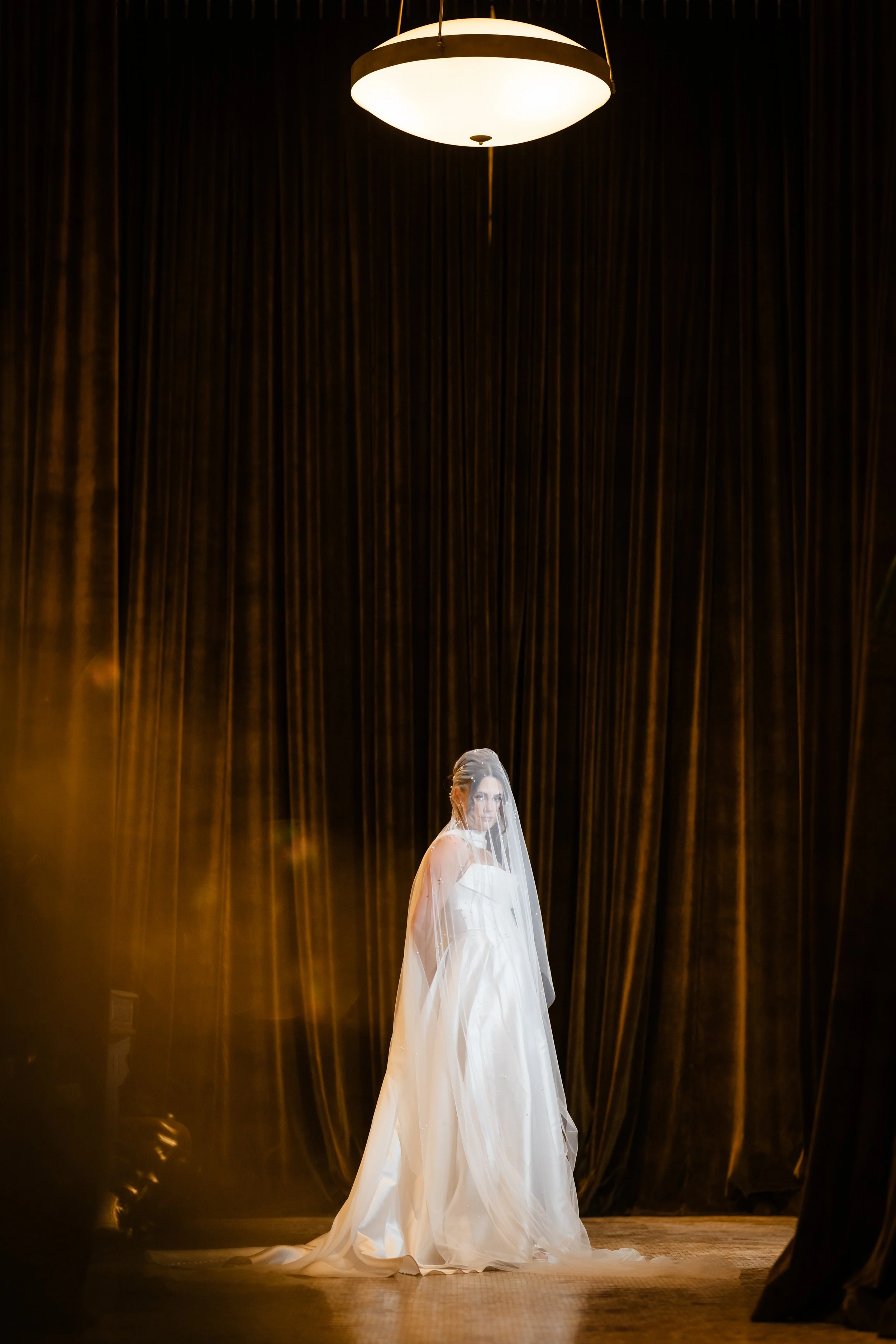 A bride in a white wedding dress with a veil standing on stage, framed by dark curtains and a hanging light fixture overhead.