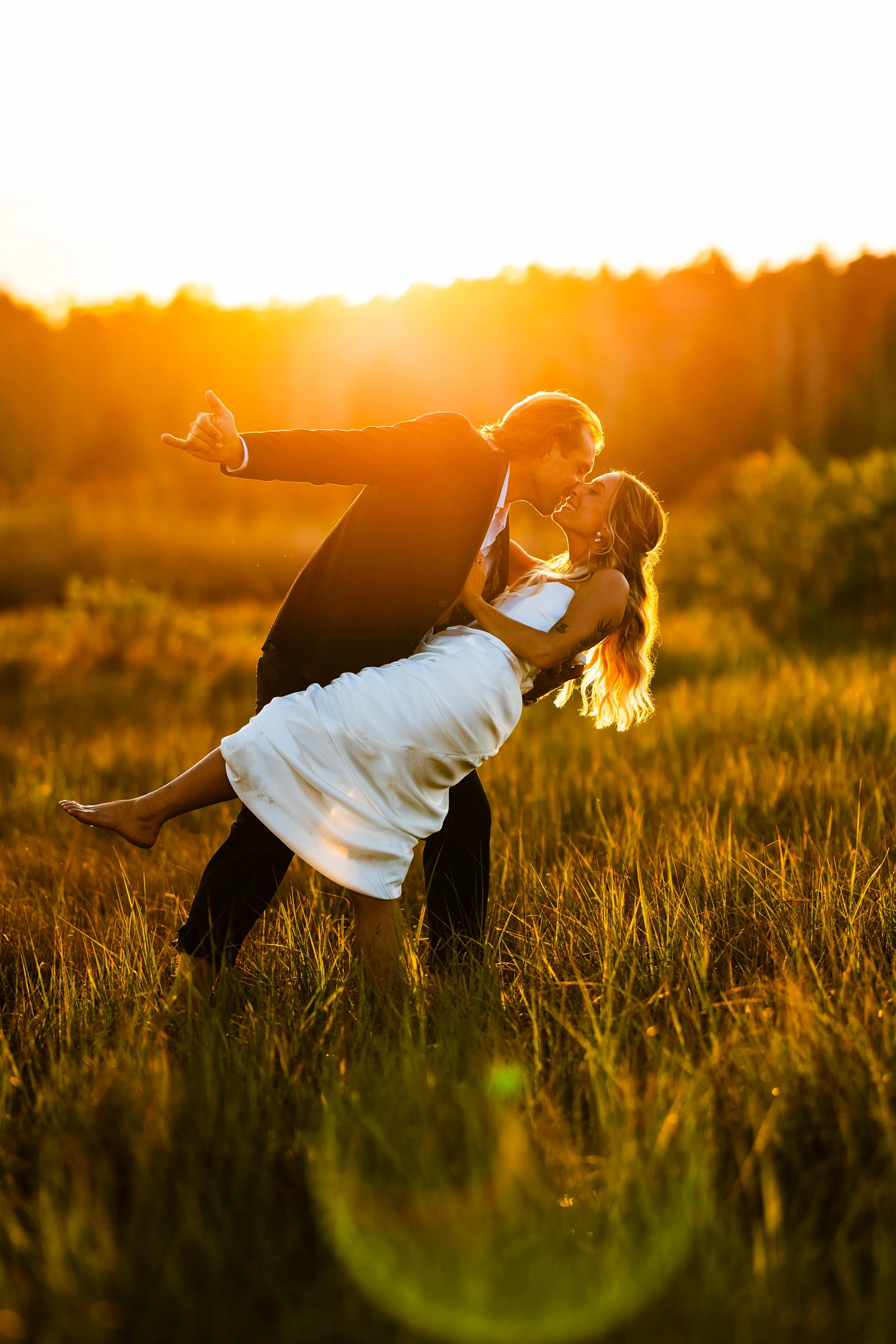 A groom in a suit dips a bride in a white dress, and they kiss during sunset in a grassy field.