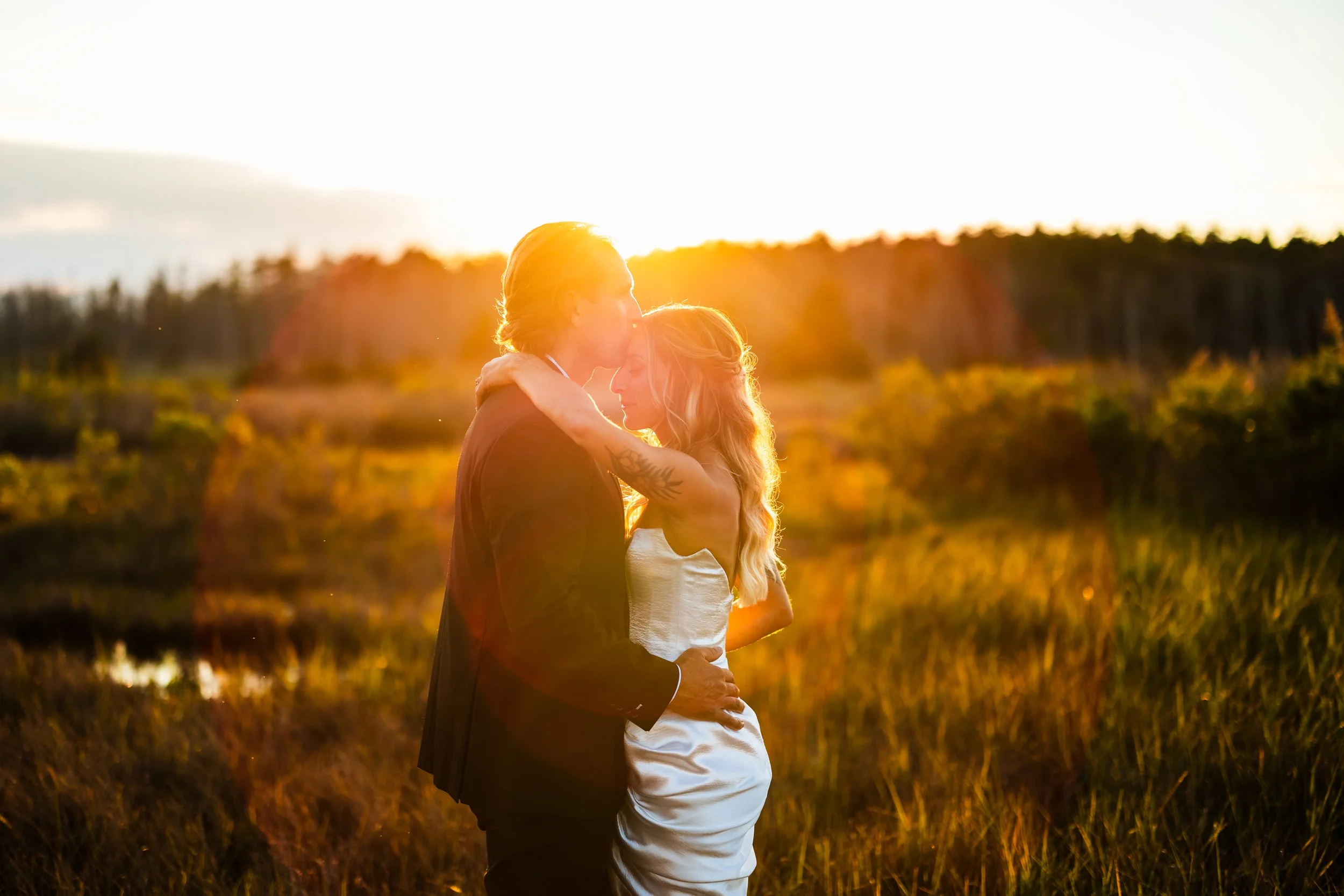A couple embracing at sunset outdoors, with golden sunlight illuminating them and a background of trees and fields.