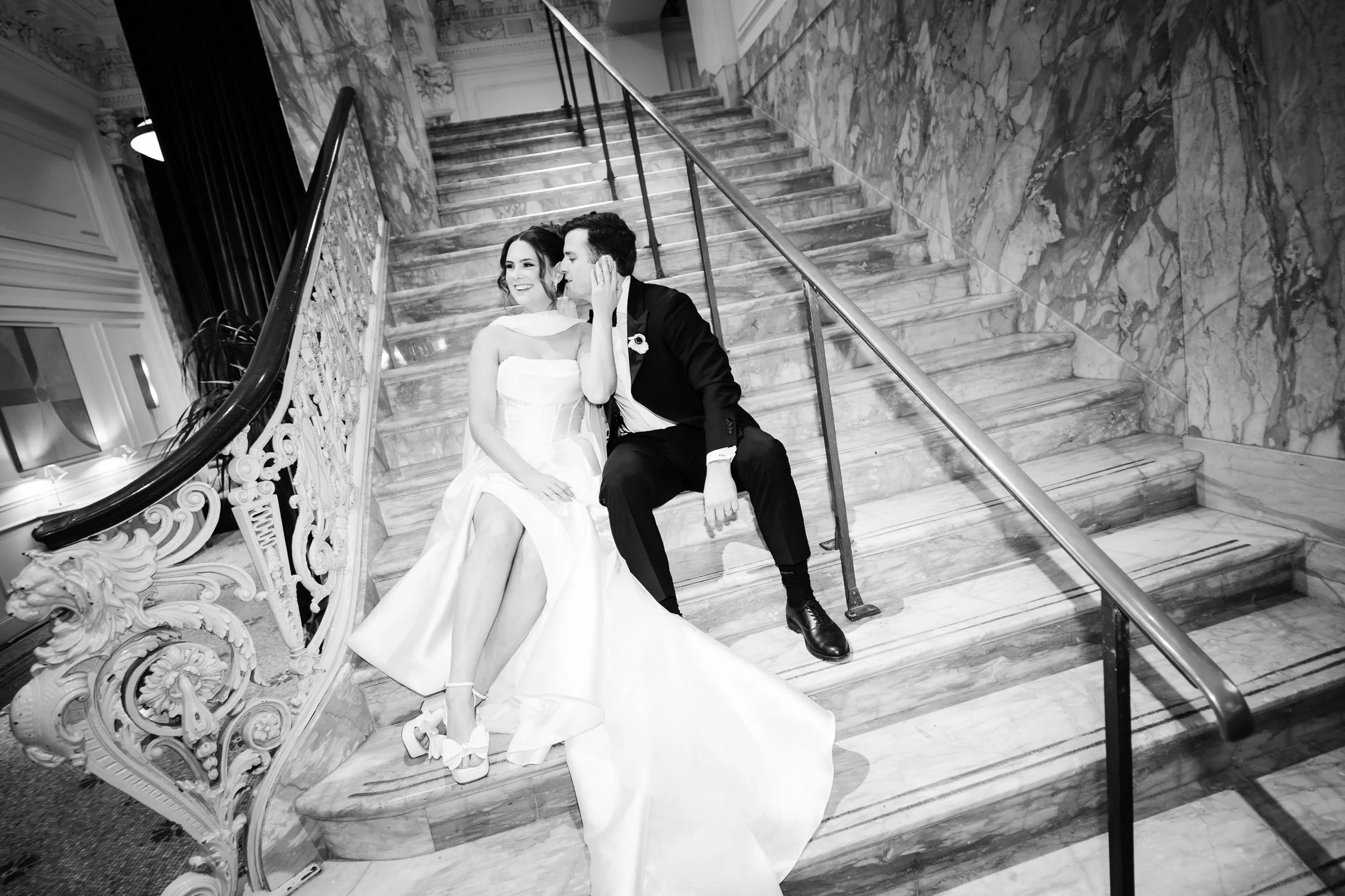 Black and white photo of a bride and groom sitting on a marble staircase, smiling and looking at each other.