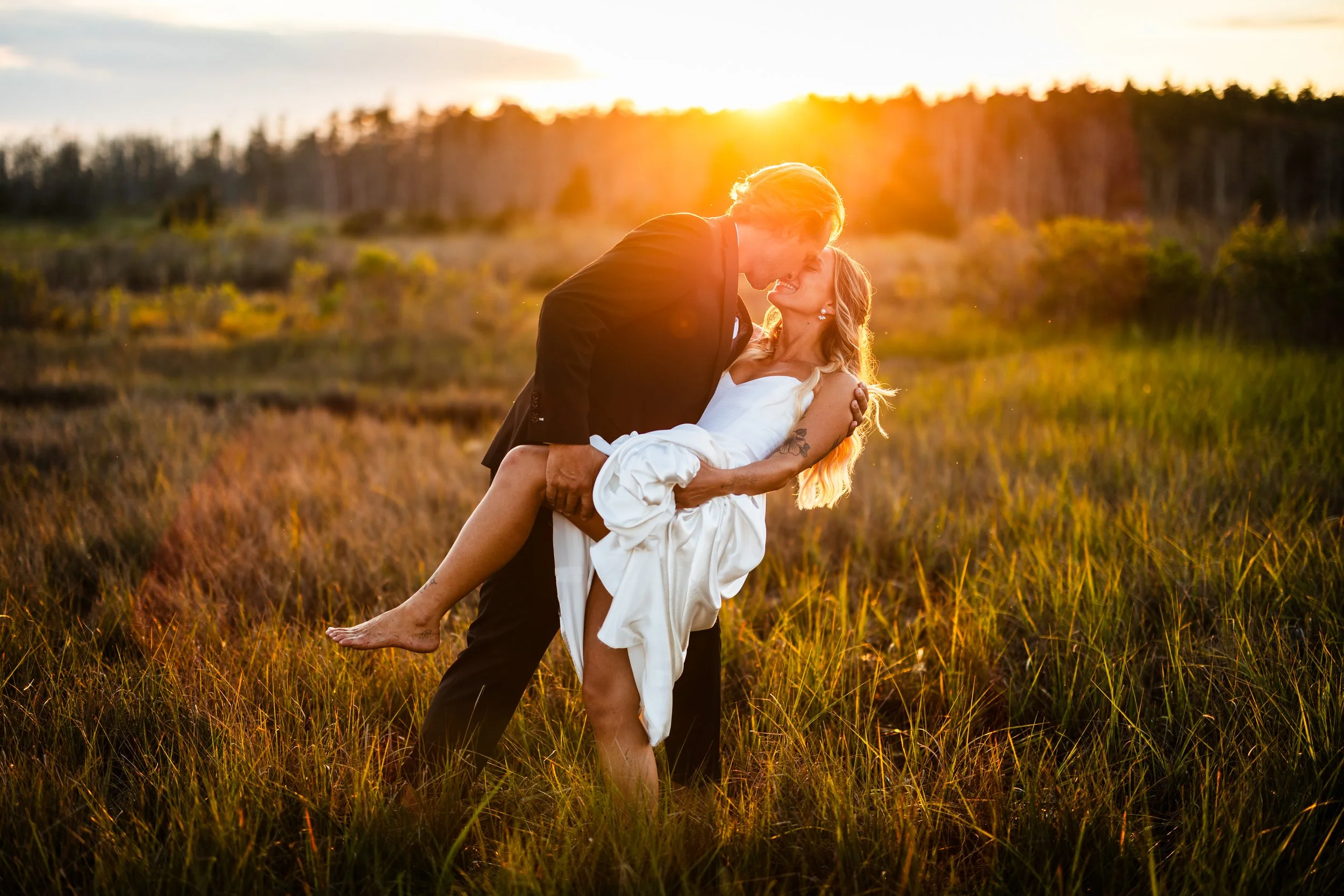 A man in a black suit holding a woman in a white dress in a field at sunset