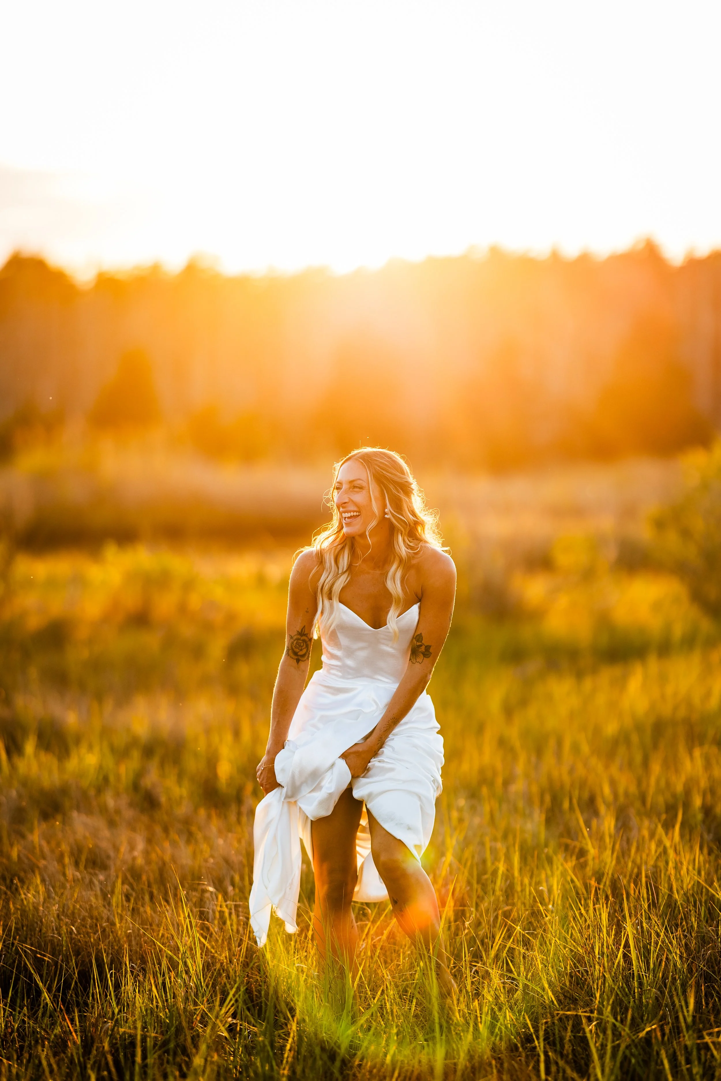 A woman in a white dress laughing and lifting her dress in a grassy field at sunset.