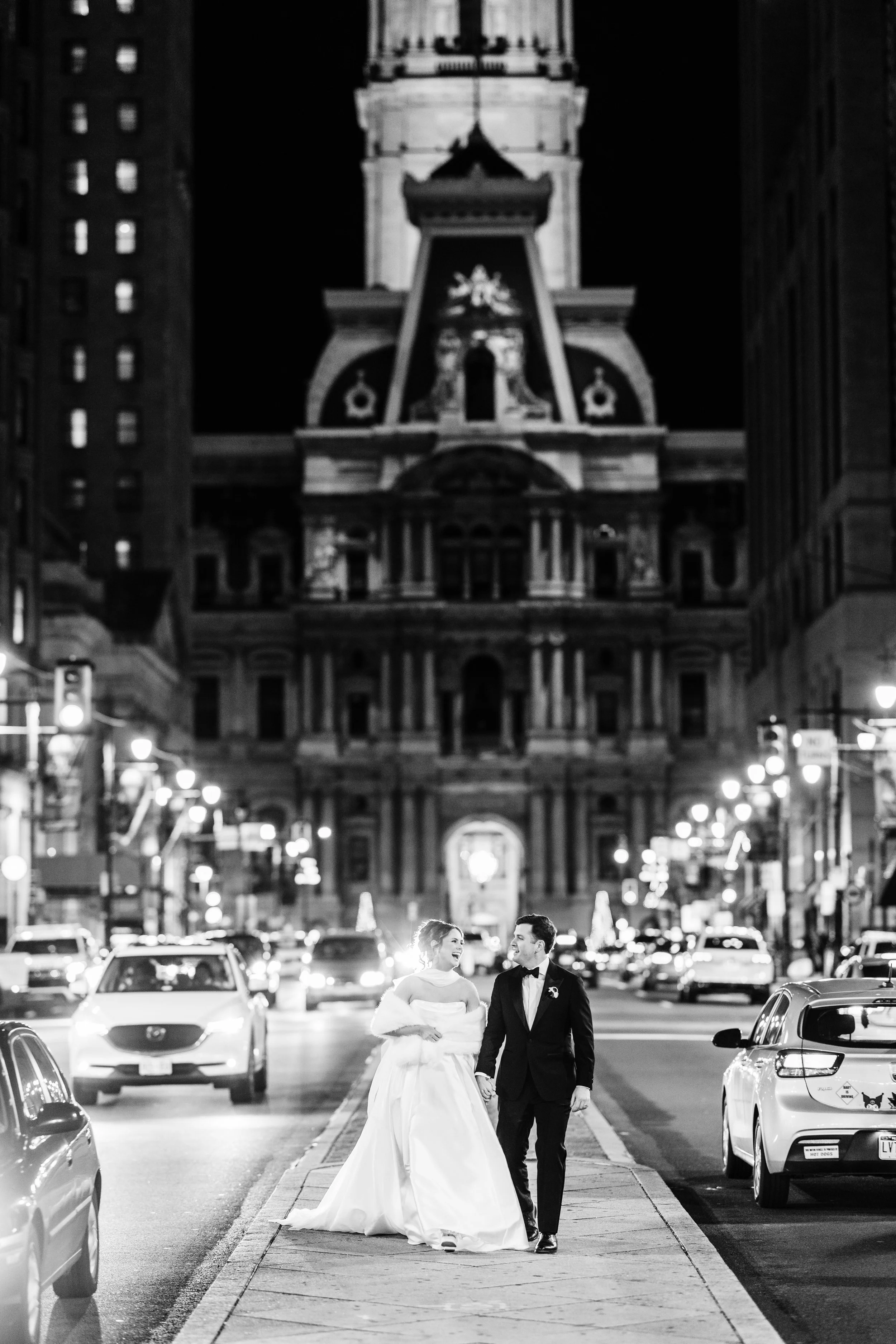 A newlywed couple in wedding attire walking and laughing on a city sidewalk at night, with a large historic building with a clock tower in the background and city lights around.