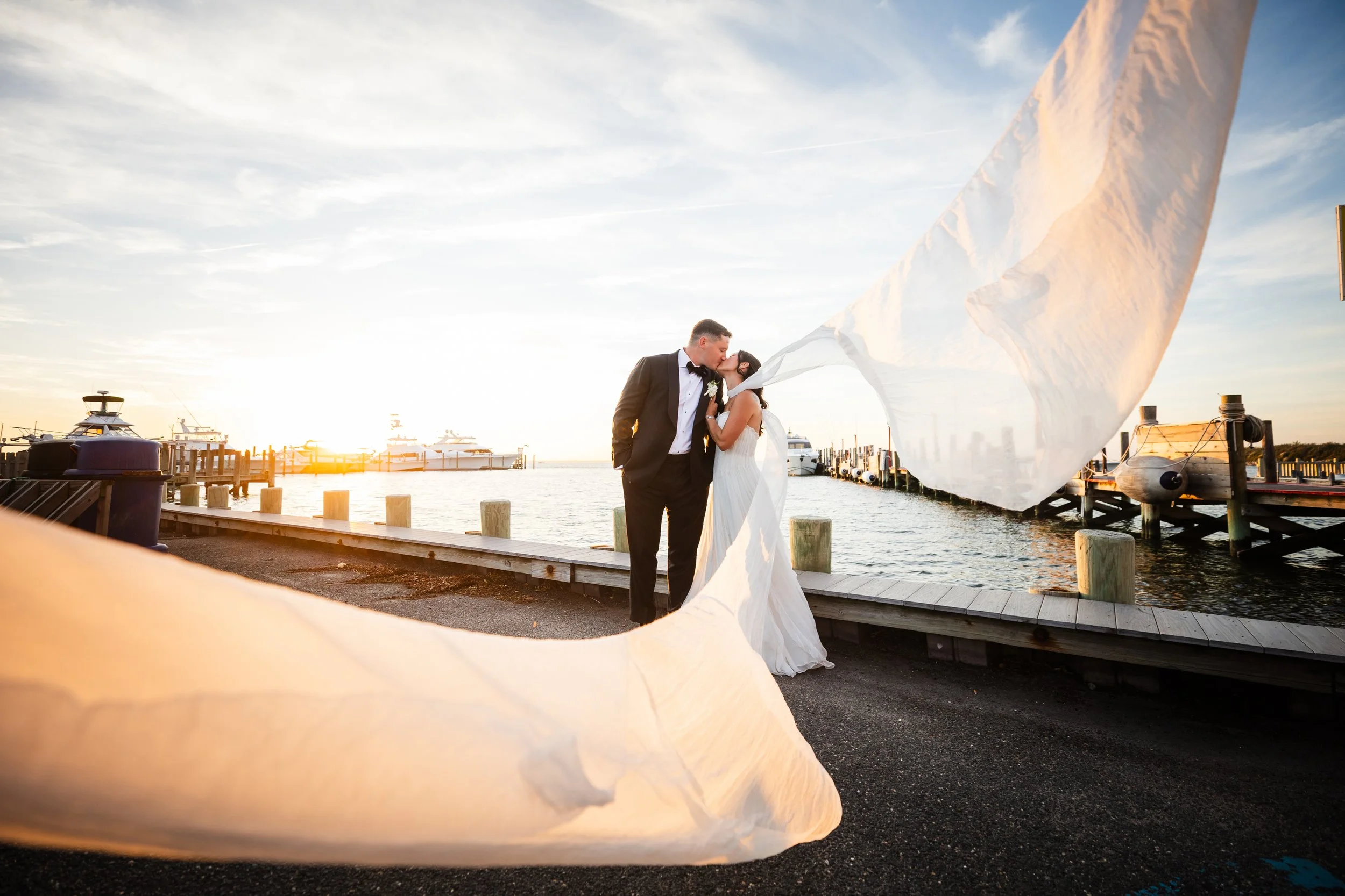 A bride and groom kissing on a dock at sunset, with their veil flowing in the wind, near boats at a harbor.