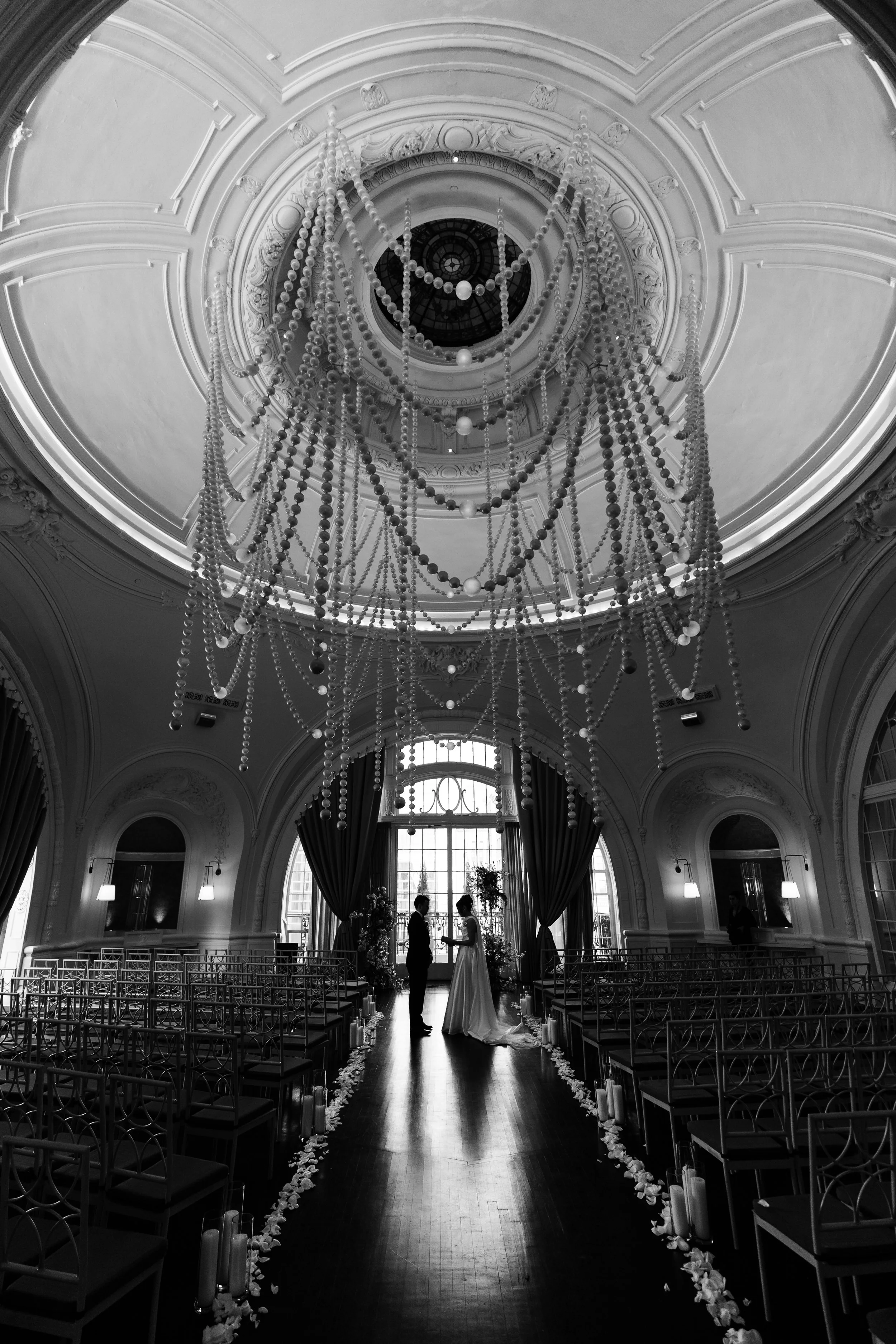 A couple getting married in an elegant, historic room with high ceilings, a large chandelier, and tall windows with curtains.